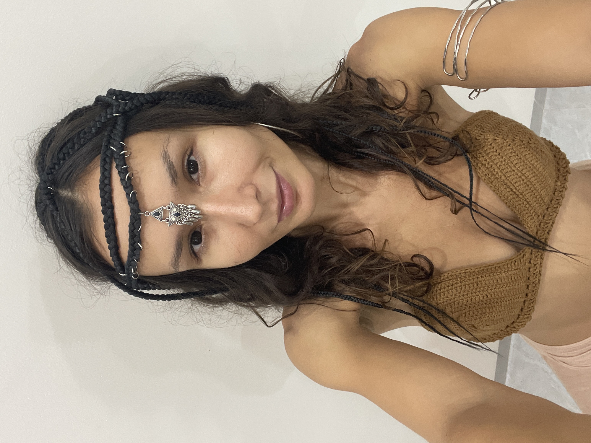 A young woman with dark, wavy hair braided with black extensions and adorned with a black braided headband featuring small silver hoops and a decorative silver pendant with a dark blue jewel, poses for a selfie. She is wearing a brown crocheted bikini top and a pale pink bottom. A silver arm cuff is visible on her right arm. The background is a plain white wall. The image does not provide any information about her location in Mũi Né, Vietnam.