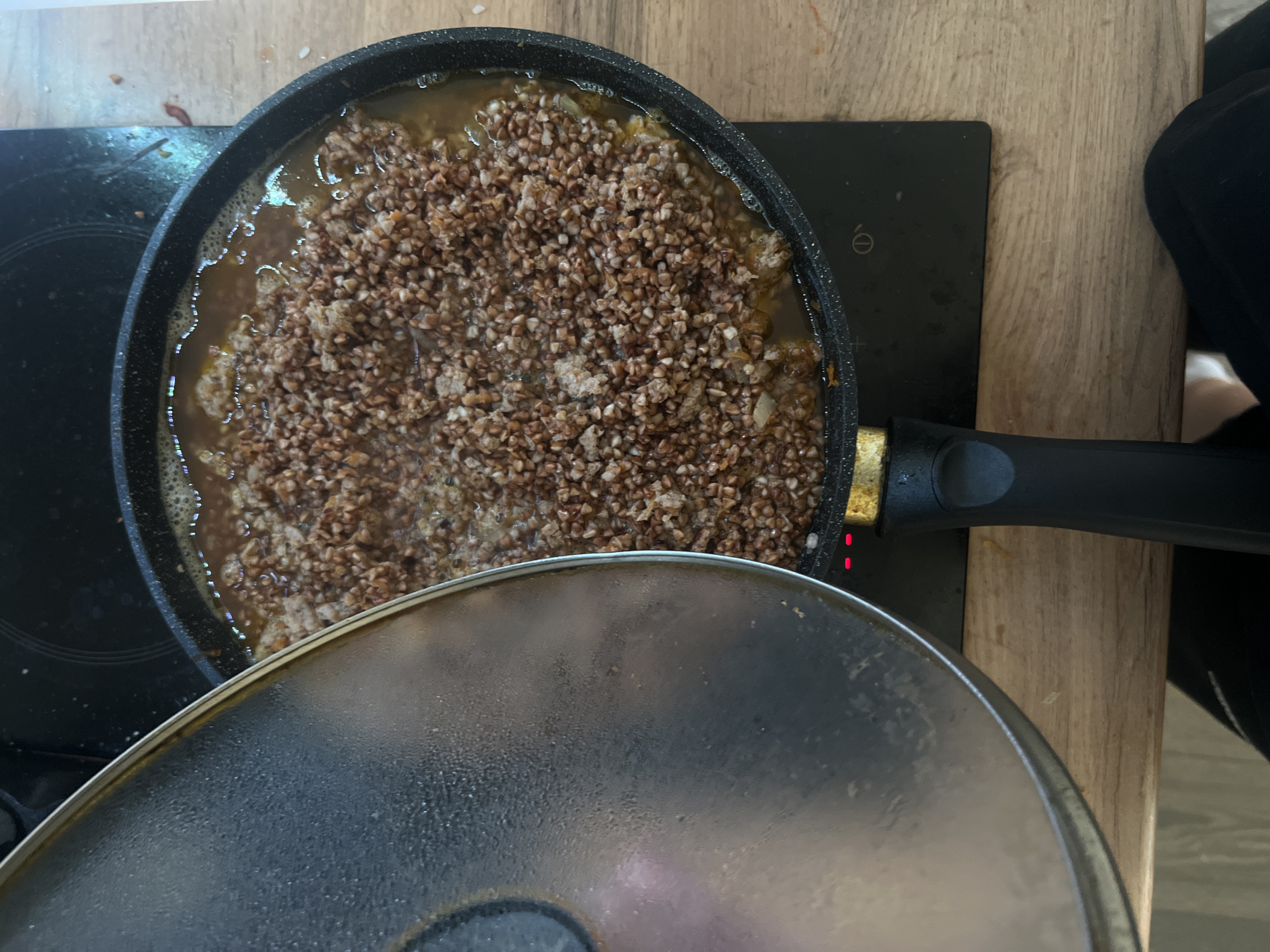 In Saint Petersburg, Russia, on a wooden countertop, a black frying pan filled with buckwheat and broth is being cooked on a black induction stove. A slightly opened metal lid is resting on the left side of the pan, showing condensation on its surface. The scene appears to be a home kitchen.