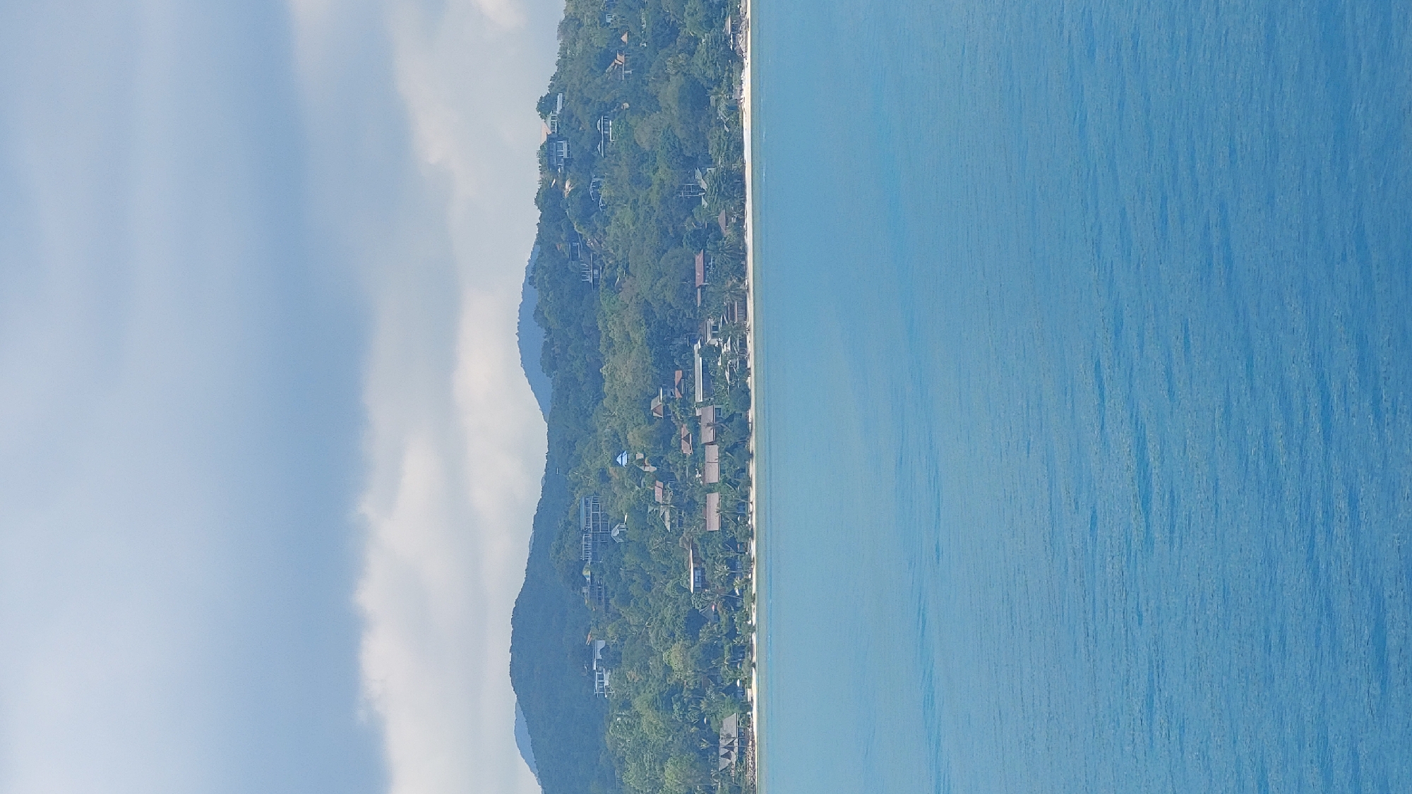 The image shows a coastal scene, likely in Baan Nai Wok, Thailand, based on the presence of lush, green vegetation on a hillside, beachside properties and the clear blue water. The scene includes a mountain covered in greenery on the left side and the calm sea on the right. There is visible evidence of sparse buildings within the trees, which indicates the area has properties. It is a sunny day, with few clouds.