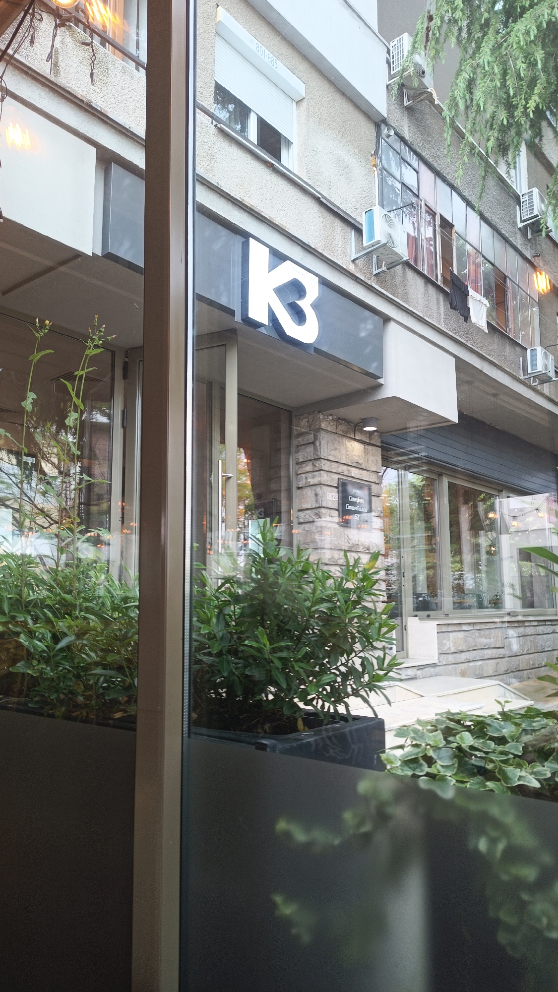 The image shows a storefront with the sign "K3" above the entrance. The location is in Burgas, Bulgaria, as indicated by the Cyrillic text on the sign next to the door, which translates to "Stefan Stambolov 52". The storefront appears to be a business, possibly a restaurant or cafe. Planters with green plants are visible in front of the glass windows, partially obscuring the view inside. The building's architecture is a mix of modern and older styles, with concrete and stone elements. Air conditioning units are visible on the building facade above the storefront. Laundry is hanging from windows in the apartment building above. The image is taken from behind a glass panel, possibly from inside another building or establishment.