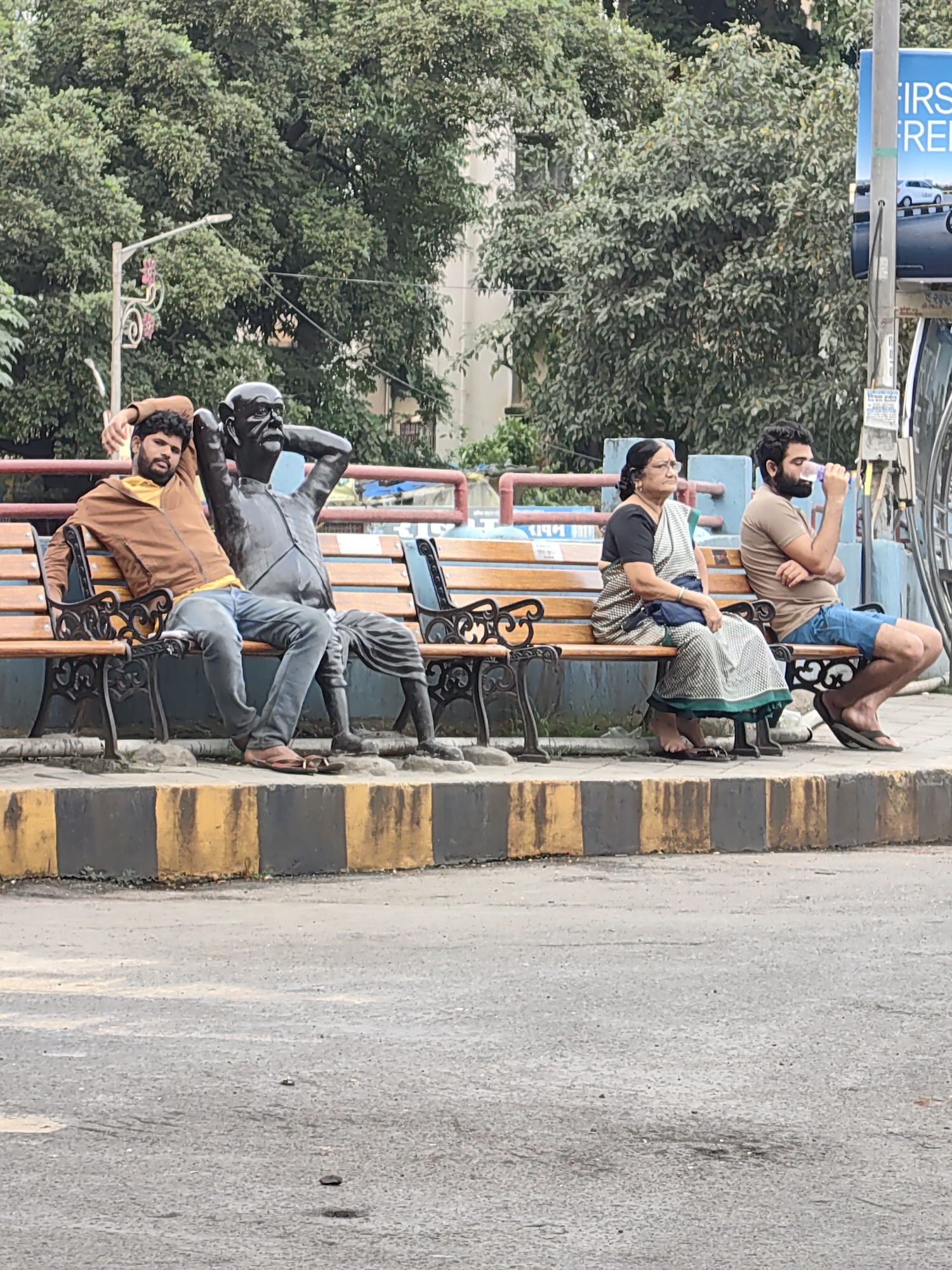 A statue of Mahatma Gandhi sits on a park bench next to a young man wearing a brown jacket and a yellow t-shirt. They are both on a wooden bench with metal legs, and the man has his arm behind his head and is looking at the camera. To the right of the statue, an elderly woman in a white and green sari is seated, looking to her left. Next to her, a man in a brown t-shirt and blue shorts drinks from a plastic bottle. The bench is located on a sidewalk with a yellow and black striped curb, and trees are visible in the background. A sign with the word "FIRST" and "FREE" in blue and white lettering is visible on the right side of the frame. The overall scene appears to be a public park or street area in Mumbai, India, given the cultural context suggested by the statue and clothing.