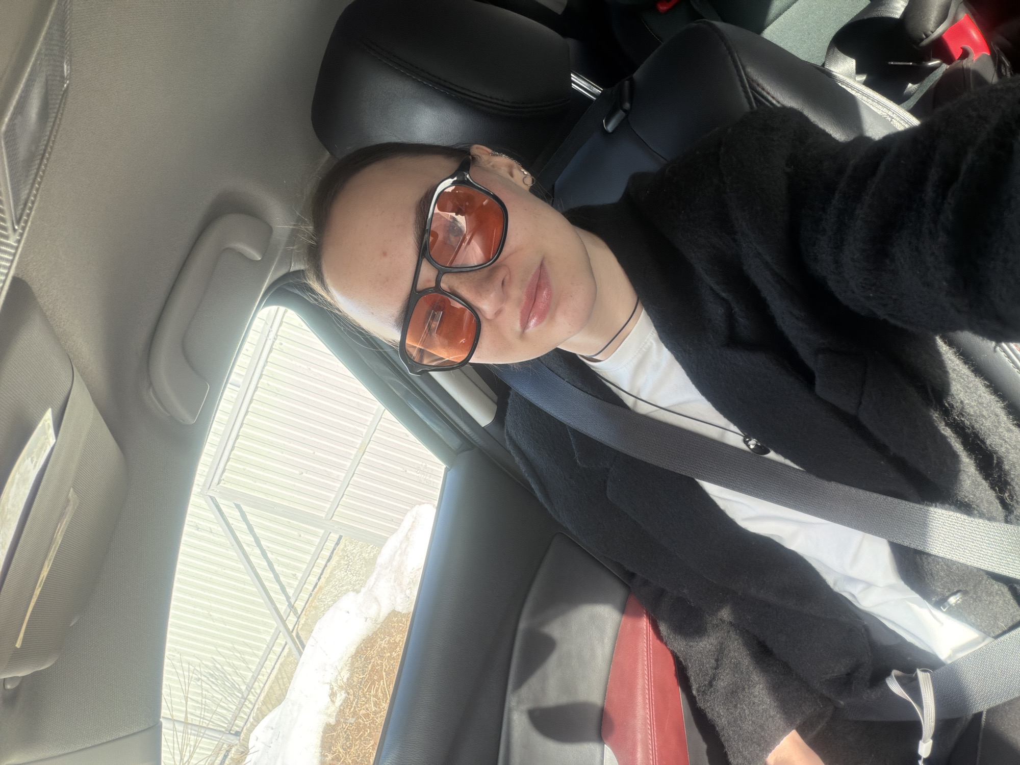 A female individual is depicted in a front-facing selfie, seated in the passenger seat of a vehicle in Chaikovsky, Russia. She wears orange-tinted, square-framed sunglasses, a white t-shirt, and a black textured jacket, secured by a grey seatbelt. Her hair is pulled back, and she has multiple ear piercings and a dark necklace with a small pendant. The vehicle's interior features black seating, a dark grey door panel with a red lower section, a light grey headliner, and a grey grab handle. Through the passenger window, patches of melting snow are visible on brown ground or sparse vegetation, backed by a light-colored, vertically corrugated metal fence or building wall under bright daylight.