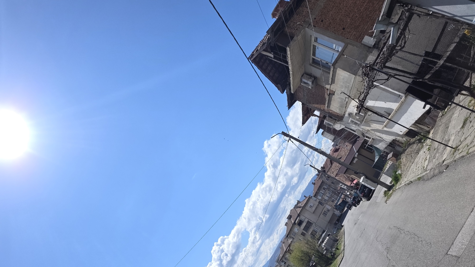 The image depicts a street in Blagoevgrad, Bulgaria, under a bright blue sky with some clouds. The sun is visible in the upper left corner. Buildings line the street, varying in architecture, with some appearing older and in need of repair. A utility pole stands near the buildings, with wires running across the sky. The street is paved and appears to be empty of pedestrians or vehicles at the immediate foreground, though vehicles are seen farther down the road.