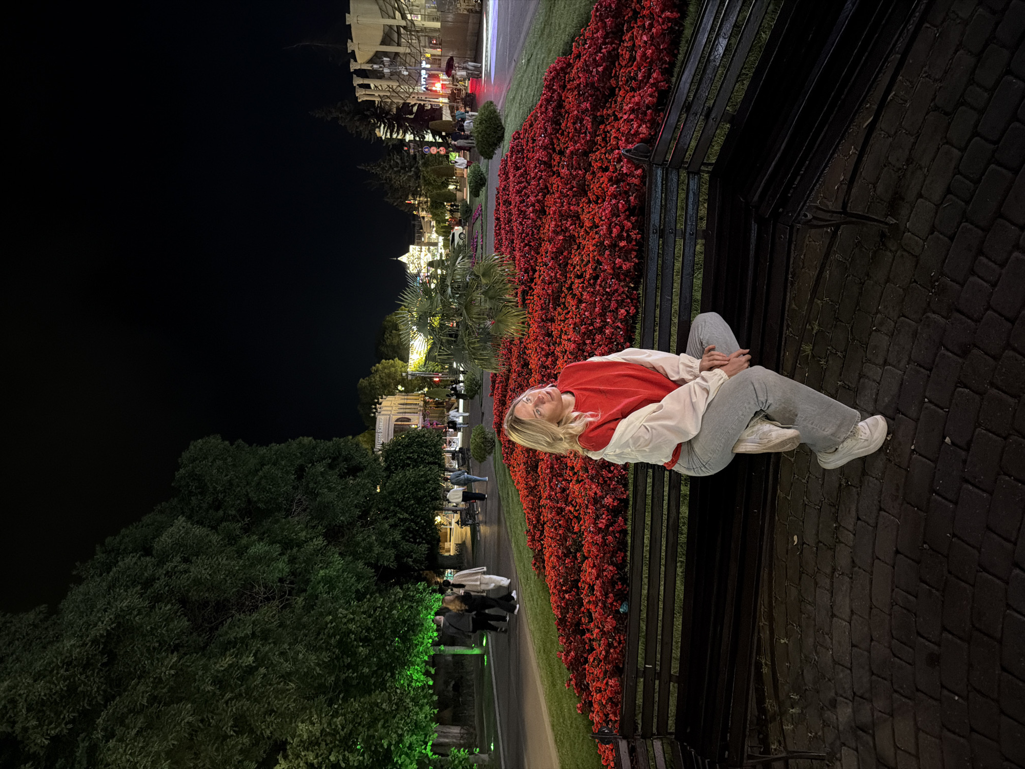A medium shot captures a young Caucasian woman with blonde hair sitting on a dark park bench at night in Kislovodsk, Russia. She is wearing a red sweatshirt with the sleeves rolled up, a light-colored jacket draped over her shoulders, light gray pants, and white sneakers. Her gaze is directed upwards and to her right, away from the camera.
The bench is situated on a paved walkway made of dark cobblestones. Behind the bench is a large, vibrant flower bed filled with numerous small red flowers, creating a striking contrast against the dark surroundings. Beyond the flower bed, a pathway is visible with several people walking in the distance. Trees line the pathway, some of which are illuminated with green lights.
In the background, the scene depicts a city street at night, with buildings illuminated by streetlights and interior lights. Architectural details of the buildings suggest a European style. The sky is dark, indicating nighttime. The overall ambiance is calm and relaxed, with the artificial lighting creating a pleasant atmosphere.
