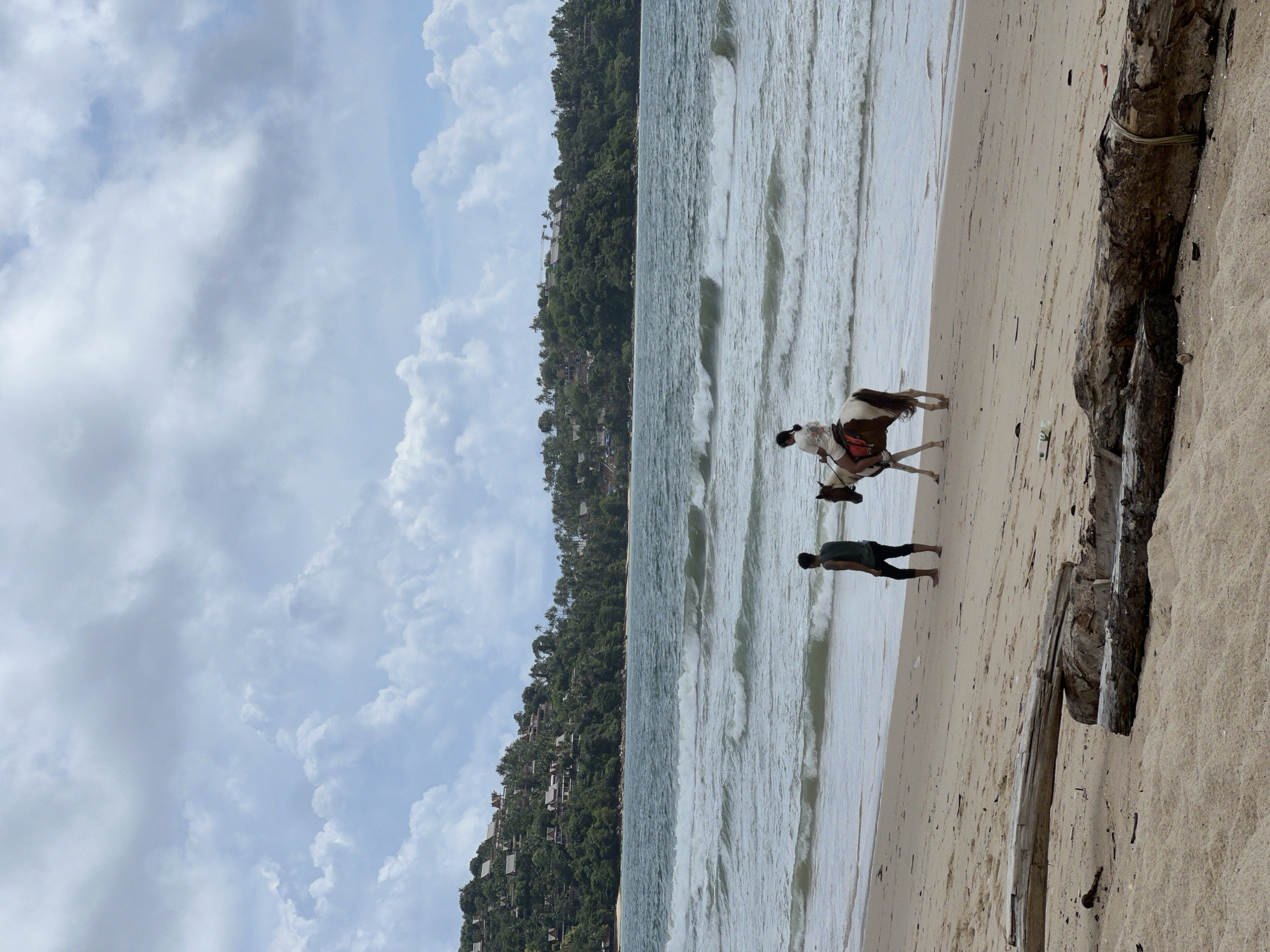 A person rides a brown and white horse along a sandy beach in Choeng Thale, Thailand, with another person walking beside them. The ocean is to the left, with moderate waves and a treed, hilly coastline in the distance. The sky is partly cloudy. In the foreground, several pieces of driftwood are scattered on the sand.