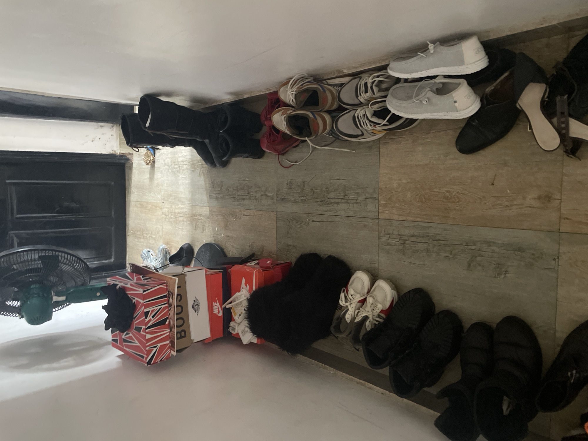 The image depicts an indoor scene with a variety of objects scattered on the floor against a wall with wood-look tiles. In the left foreground, there is a green oscillating fan. To the right of the fan, shoe boxes are stacked. An assortment of shoes, including sneakers, sandals, and boots, are arranged along the base of the wall, some in pairs and some individually. The shoes are in varying conditions and styles. The scene is indoors, likely a domestic entryway. The city and country were provided, but the image itself does not provide sufficient information to verify this.