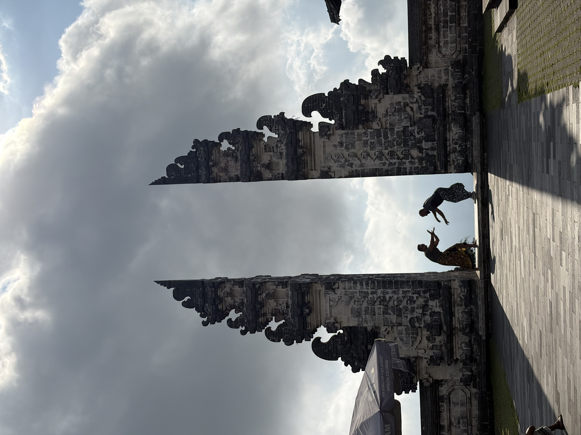 The image depicts the iconic split gate at the Lempuyang Temple in Abang, Indonesia, famously known as the "Gates of Heaven." Two individuals, silhouetted against the sky, are positioned in the center of the frame, between the two towering stone structures of the gate. They appear to be interacting, with one person reaching out towards the other, possibly in a playful or performative gesture. The gate itself is ornately carved and weathered, showcasing traditional Balinese architecture. The background is dominated by a dramatic sky filled with large, textured clouds, some illuminated by sunlight, suggesting a dynamic weather condition. In the foreground, a paved walkway with a distinctive pattern of light and shadow stretches towards the gate. To the left, a dark umbrella with text, including "FIFA WORLD CUP Qatar2022" and "OFFICIAL ICE CREAM," is visible, indicating the presence of some commercial activity. The overall scene captures a moment of human interaction within a significant cultural and architectural landmark against a striking natural backdrop.