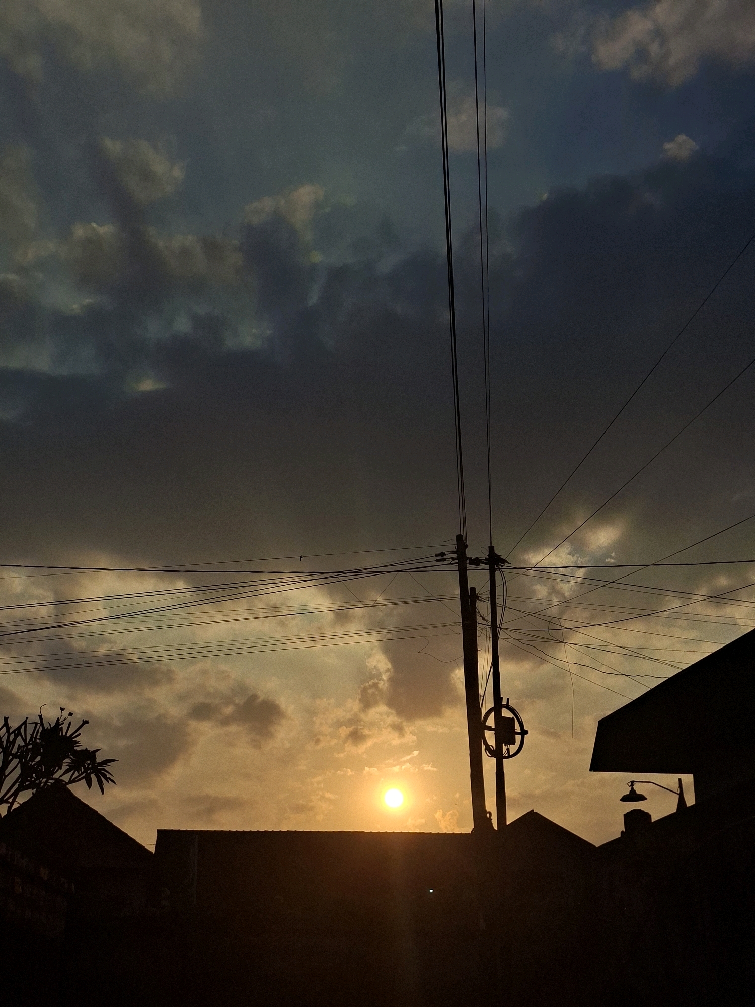 The image captures a sunset scene in Blahbatuh, Indonesia. Silhouetted against the golden sky are the rooftops of houses and a prominent utility pole with a complex network of wires. The sun, a bright orb, is partially obscured by clouds and appears low on the horizon. The sky transitions from a deep blue at the top to a warm orange and yellow near the sun. The overall atmosphere is dusky and serene.