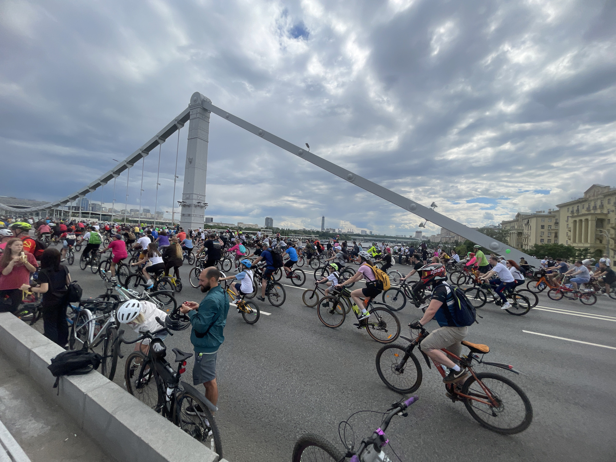 A large group of cyclists ride across a bridge in Unknown, Unknown. The bridge has a distinctive white tower and angled support structure. The sky overhead is cloudy. Many cyclists wear helmets and a variety of casual clothing. Some people are standing on the edge of the bridge, near parked bicycles. Buildings are visible in the background behind the bridge.