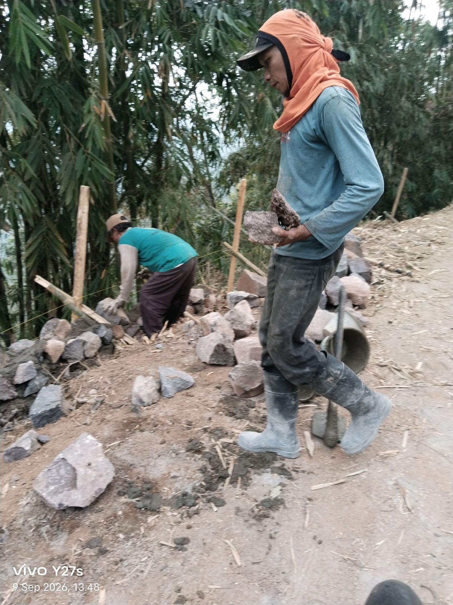 In the city of Tamansari, Indonesia, two workers are engaged in construction. In the foreground, a man wearing a blue long-sleeved shirt, dark pants, black rubber boots, and an orange head covering is walking, carrying a rock. To his left, a second worker, dressed in a teal shirt, brown pants, and a beige cap, is bent over, placing stones to build a wall. The environment is a dirt construction site with various rocks scattered around, bamboo trees in the background, and visible soil and debris. The image is dated September 9, 2026, at 13:48.
