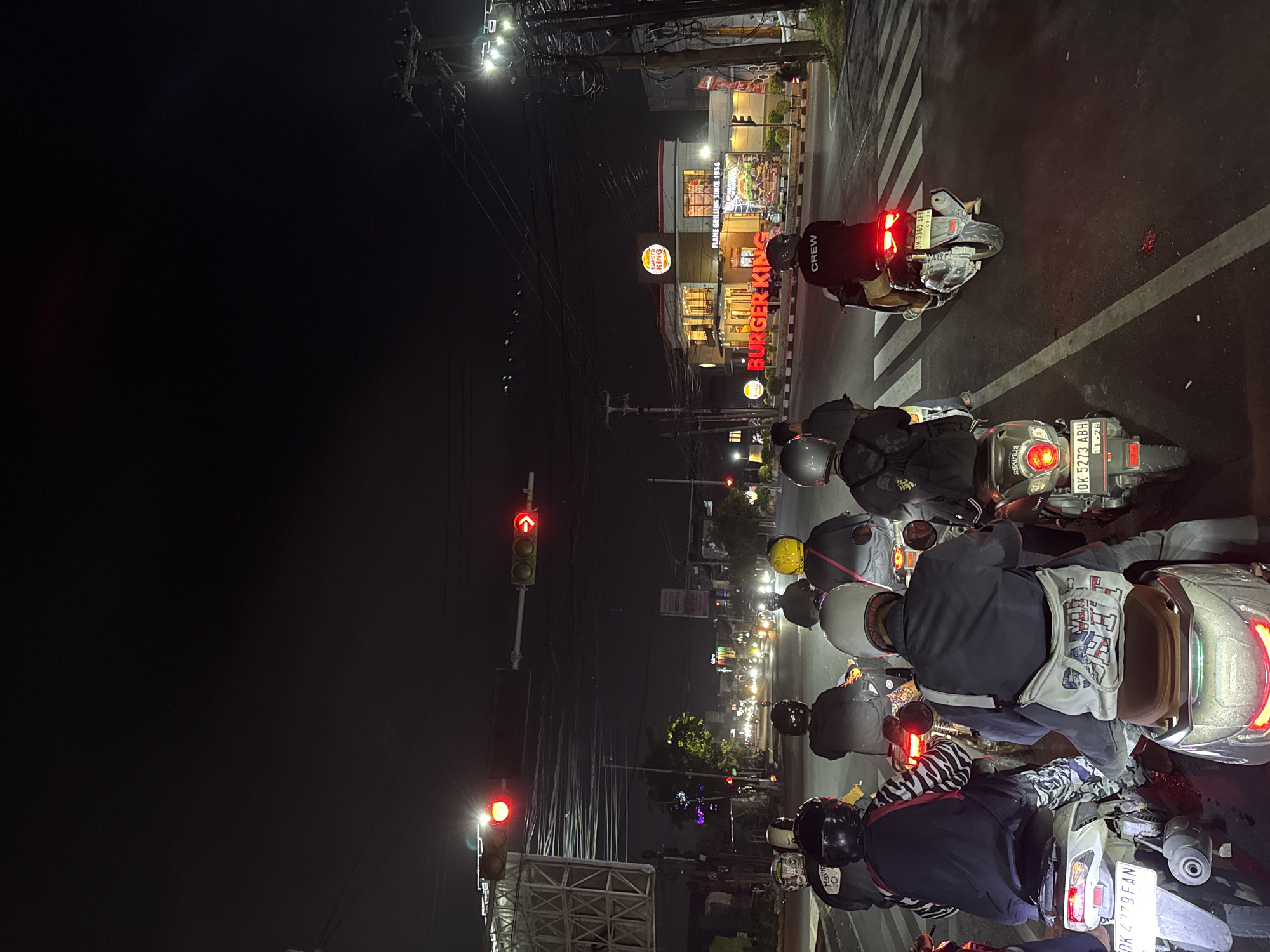 The image captures a nighttime street scene in Denpasar, Indonesia, likely at an intersection. A group of motorcyclists is stopped at a red traffic light, their vehicles forming a queue. The prominent Burger King establishment with illuminated signage is visible on the right side of the road, indicating a commercial area. The sky is dark, and streetlights and the lights from buildings provide illumination. The road surface appears wet, suggesting recent rain. The atmosphere is busy, with traffic lights controlling the flow of vehicles at the intersection. Several individuals are wearing helmets and casual attire. The overall scene depicts a common urban scenario in Indonesia at night.