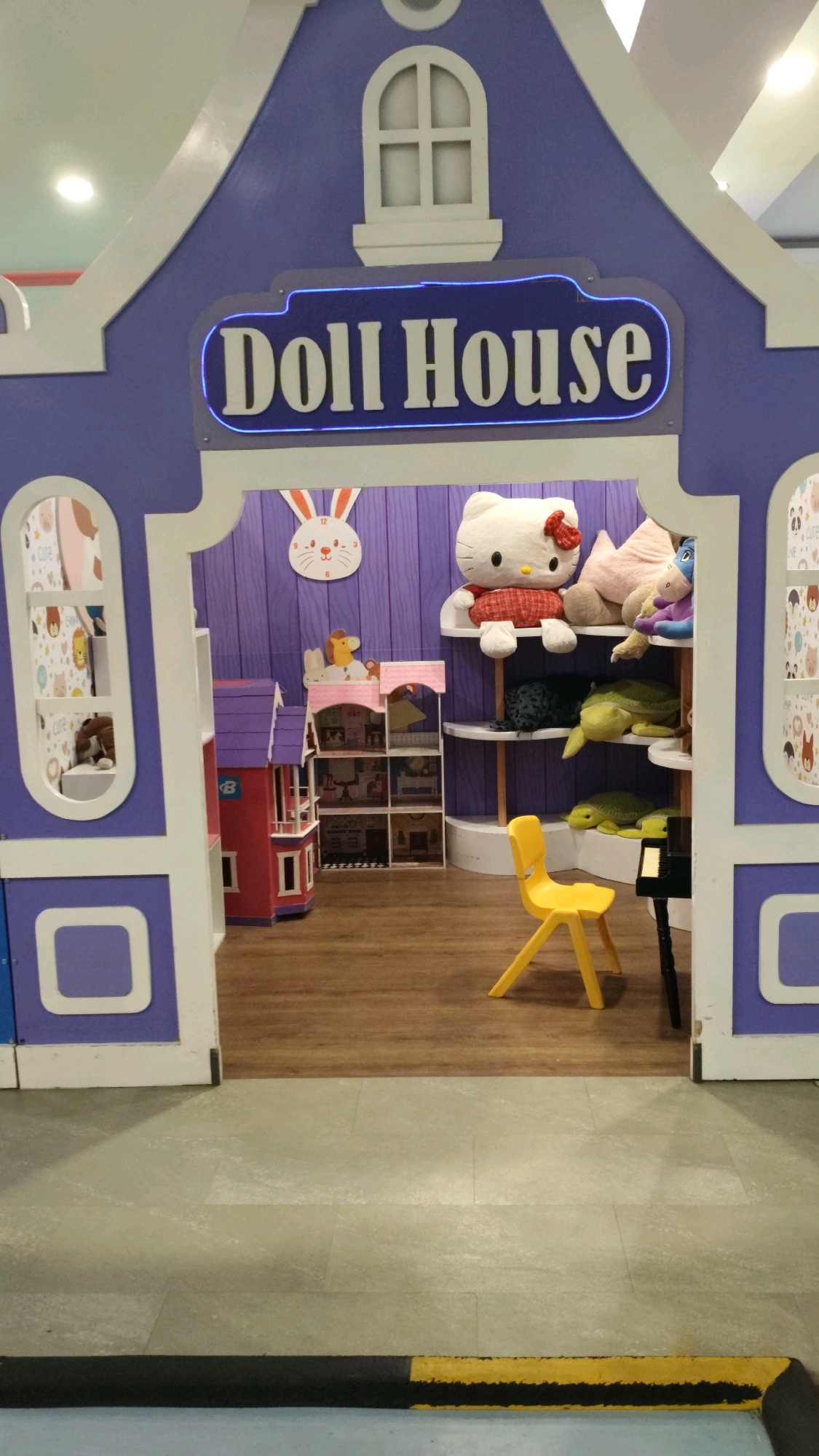 An indoor, child-friendly play area designed as a "Doll House" is depicted. The entrance features a large, purple, house-shaped structure with white trim. A neon blue sign above the entrance reads "Doll House". Inside, a purple, vertically paneled wall serves as a backdrop. On shelves, a large Hello Kitty plush toy, a pink pillow, and other stuffed animals are visible. To the left, a multi-story dollhouse is situated. A small, red and pink dollhouse accessory unit with multiple compartments is placed next to it. A bright yellow chair stands in the foreground on a wooden floor. To the right, a black object, possibly a small piano, is partially visible. The floor transitions from wood to a lighter tiled material near the entrance. The scene suggests a playful and imaginative environment, likely within a commercial establishment such as a toy store, playground, or children's entertainment center in Bandung City, Indonesia.