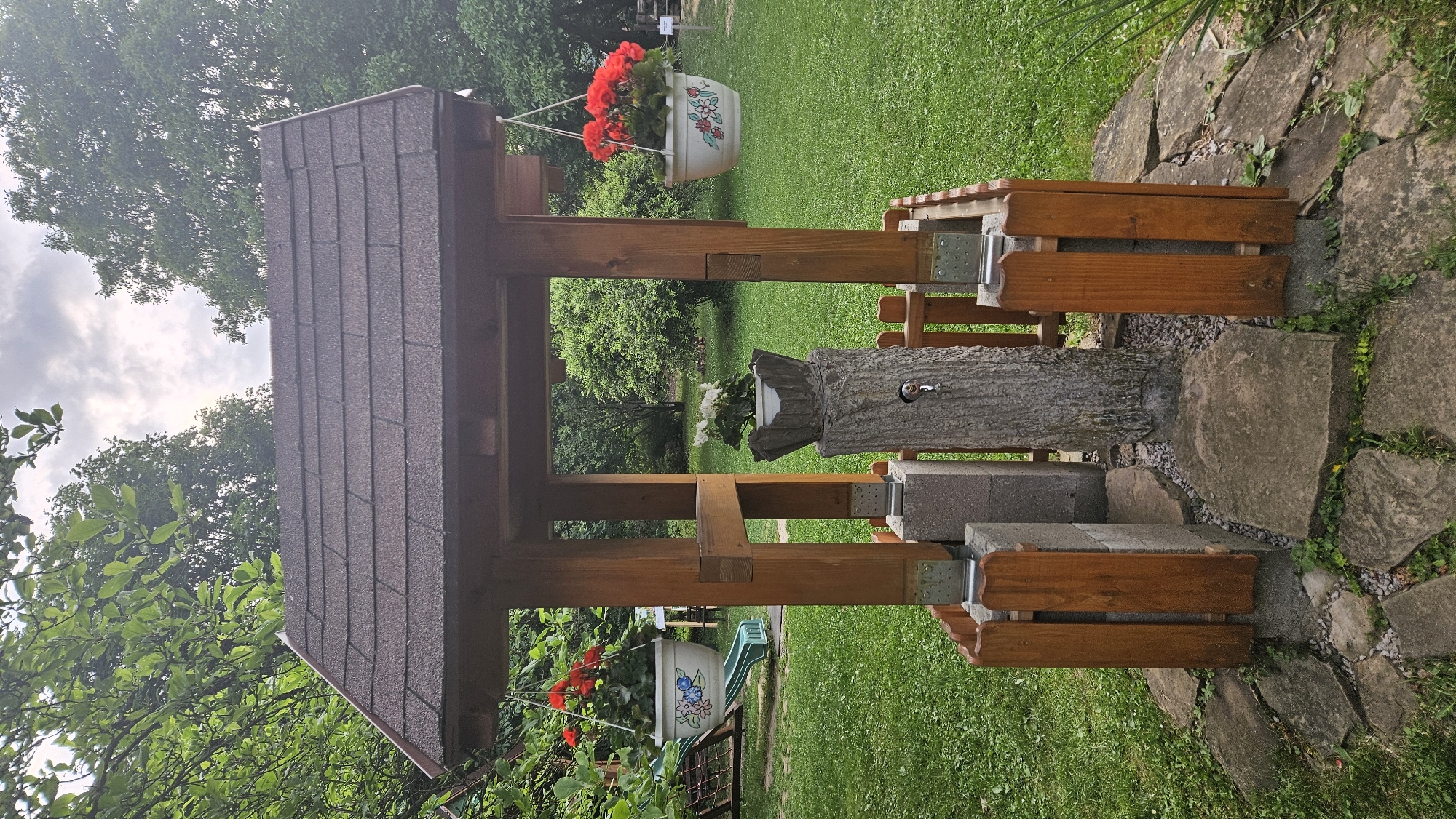 The image depicts a decorative garden well structure located in what appears to be a residential garden. The well has a dark brown shingled roof supported by four wooden posts. Hanging flower pots with red geraniums adorn two of the posts. A simulated tree trunk with a faucet protrudes from the center of the well. The structure is built on a foundation of grey cinder blocks and capped with wooden planks. The surrounding ground cover consists of grass and paving stones. The background shows greenery of trees and shrubbery. The photo was most likely taken in the city of Prešov, Slovakia.