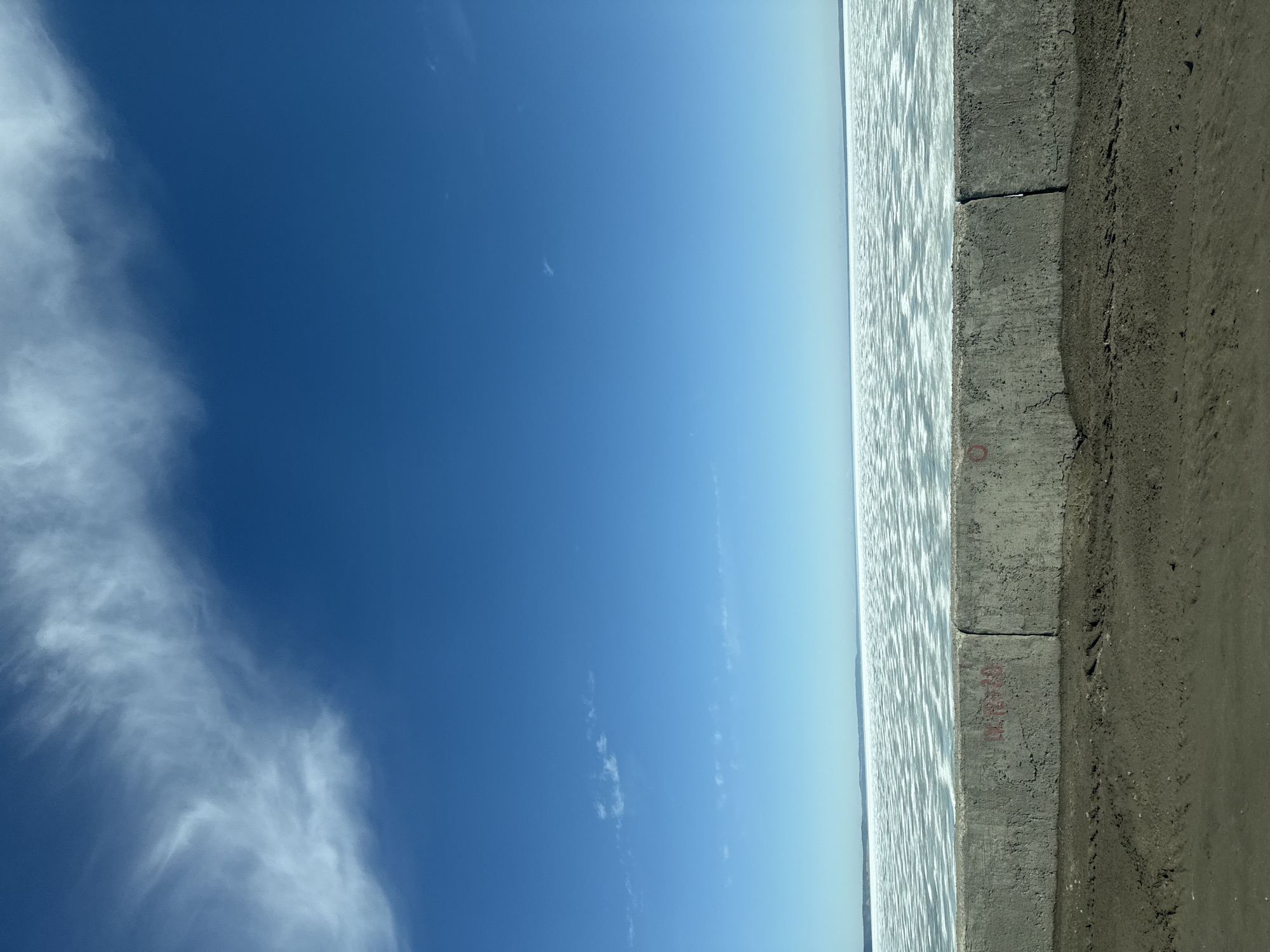 The image depicts a concrete embankment separating a body of water from a sandy shore, with a clear blue sky above. The water's surface is slightly rippled, reflecting light. The embankment consists of concrete blocks, with some minor wear and markings. The sandy shore is dark. The sky is clear with some scattered cirrus clouds on the left side. There's no indication of any people or specific actions in the frame.