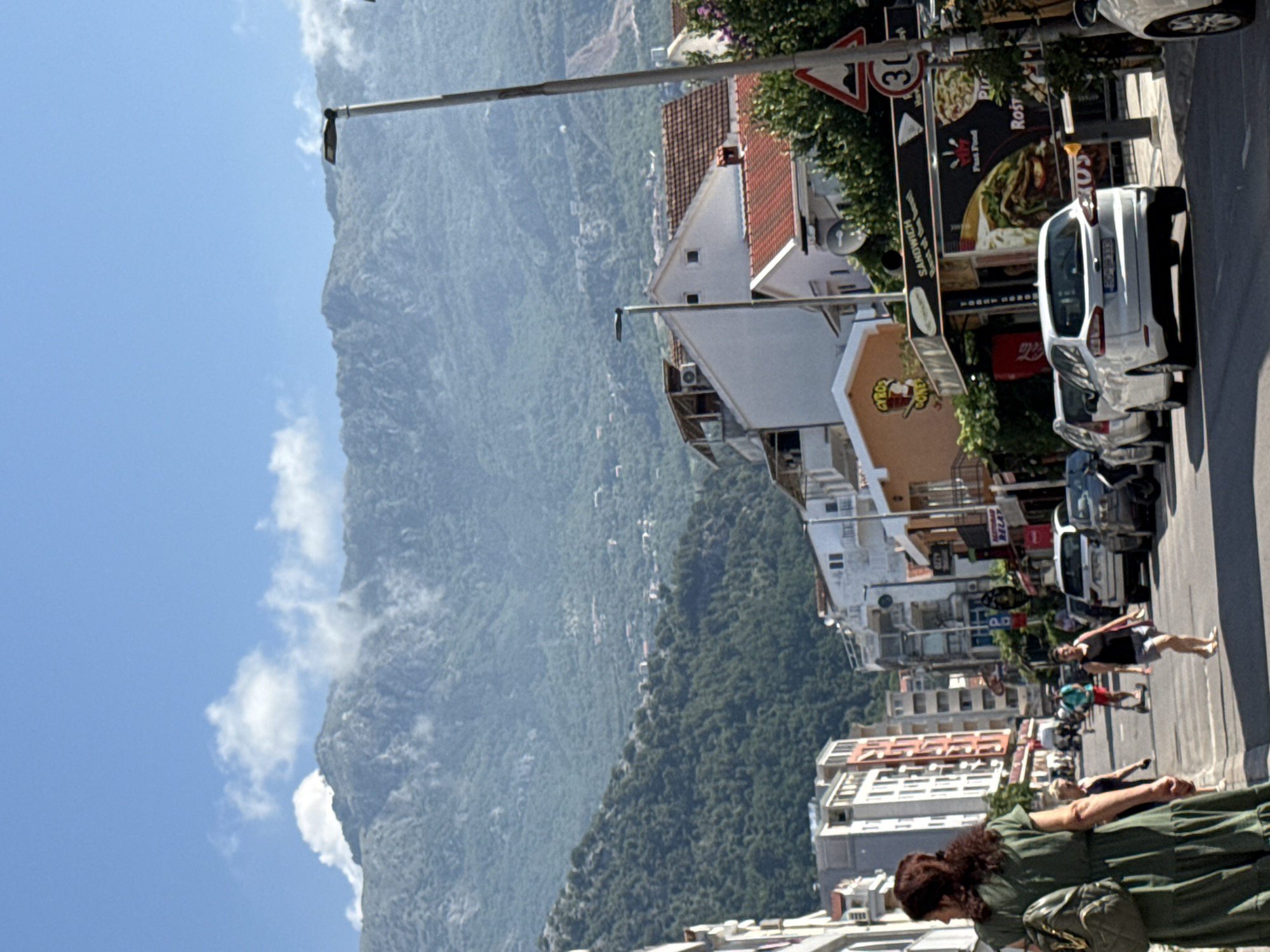 This is a street-level, eye-level, medium shot taken on a sunny day, looking up a street toward a mountainous backdrop in Budva, Montenegro. In the foreground, a woman wearing a green dress and a backpack walks away from the camera. Other people are visible walking and riding scooters on the street, with cars parked on the right side. The buildings lining the street are of various heights and architectural styles, with shops and restaurants on the ground floors. A traffic sign indicating a speed limit of 30 is visible. The background is dominated by a lush green mountain range under a blue sky with scattered clouds.