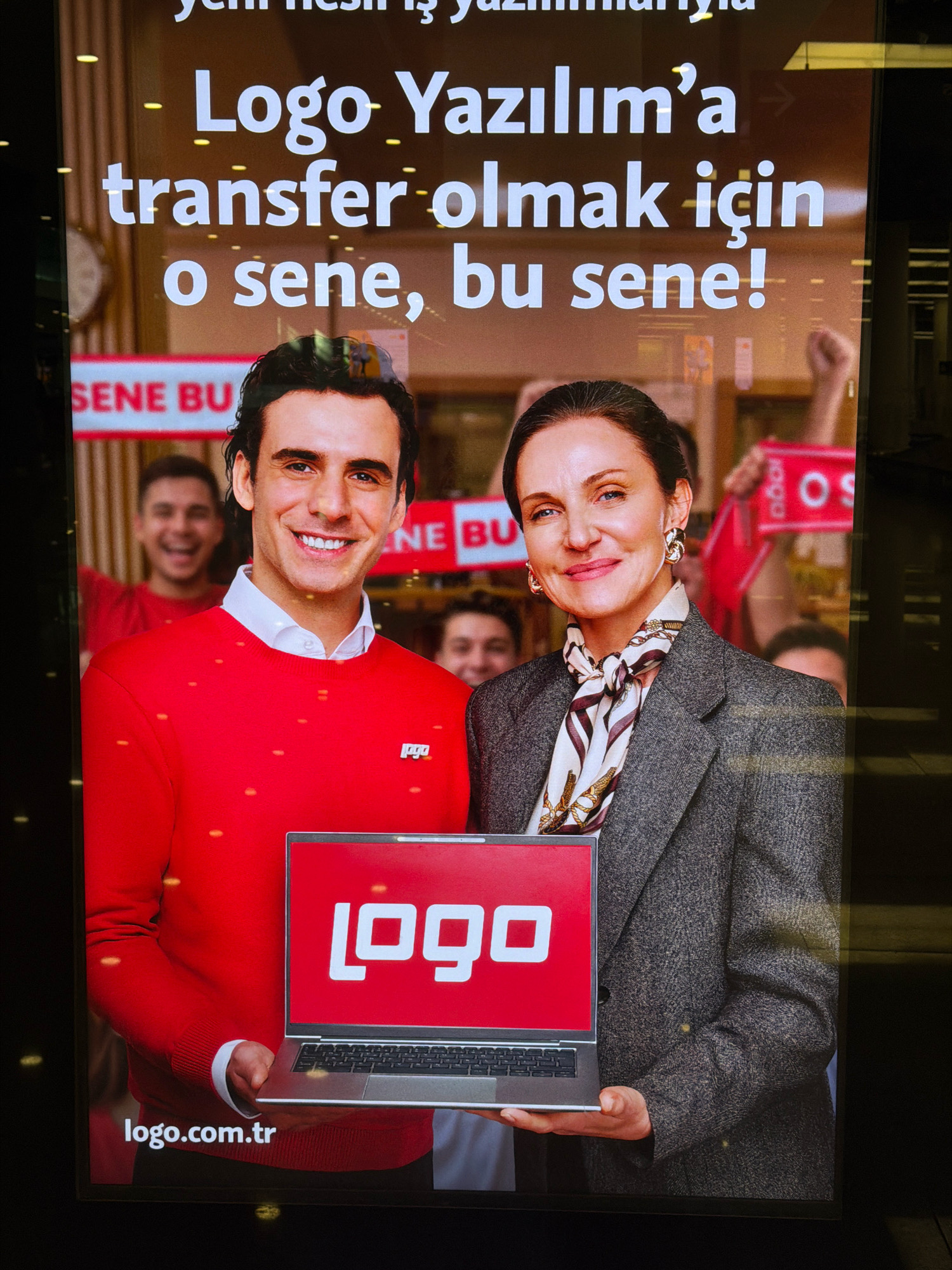 The image is a photograph of a large digital advertisement displayed on a screen, likely in an indoor public space. The advertisement features two individuals, a man and a woman, prominently in the foreground. The man, on the left, is smiling and wearing a red sweater with small orange dots. He is holding a silver laptop with a red screen displaying a white logo that reads "logo." The woman, on the right, is also looking towards the viewer with a slight smile. She is wearing a grey blazer over a patterned scarf and large gold earrings.
Behind them, other people can be seen, some holding red signs with white lettering that appears to be "SENE BU" and "OS." The background of the advertisement seems to be a stylized image of people cheering or celebrating. The text on the advertisement is in Turkish and reads, "- Logo Yazılım'a transfer olmak için o sene, bu sene!" which translates to "For transferring to Logo Software, that year, this year!" The website address "logo.com.tr" is visible at the bottom left corner of the screen. The overall impression is of a recruitment or promotional advertisement for the company Logo Yazılım.
While the image itself does not provide specific clues to its exact location within Gaziemir, Turkey, such digital advertisements are commonly found in shopping malls, airports, or busy commercial areas in cities. The lighting suggests an indoor setting, possibly a brightly lit commercial environment.