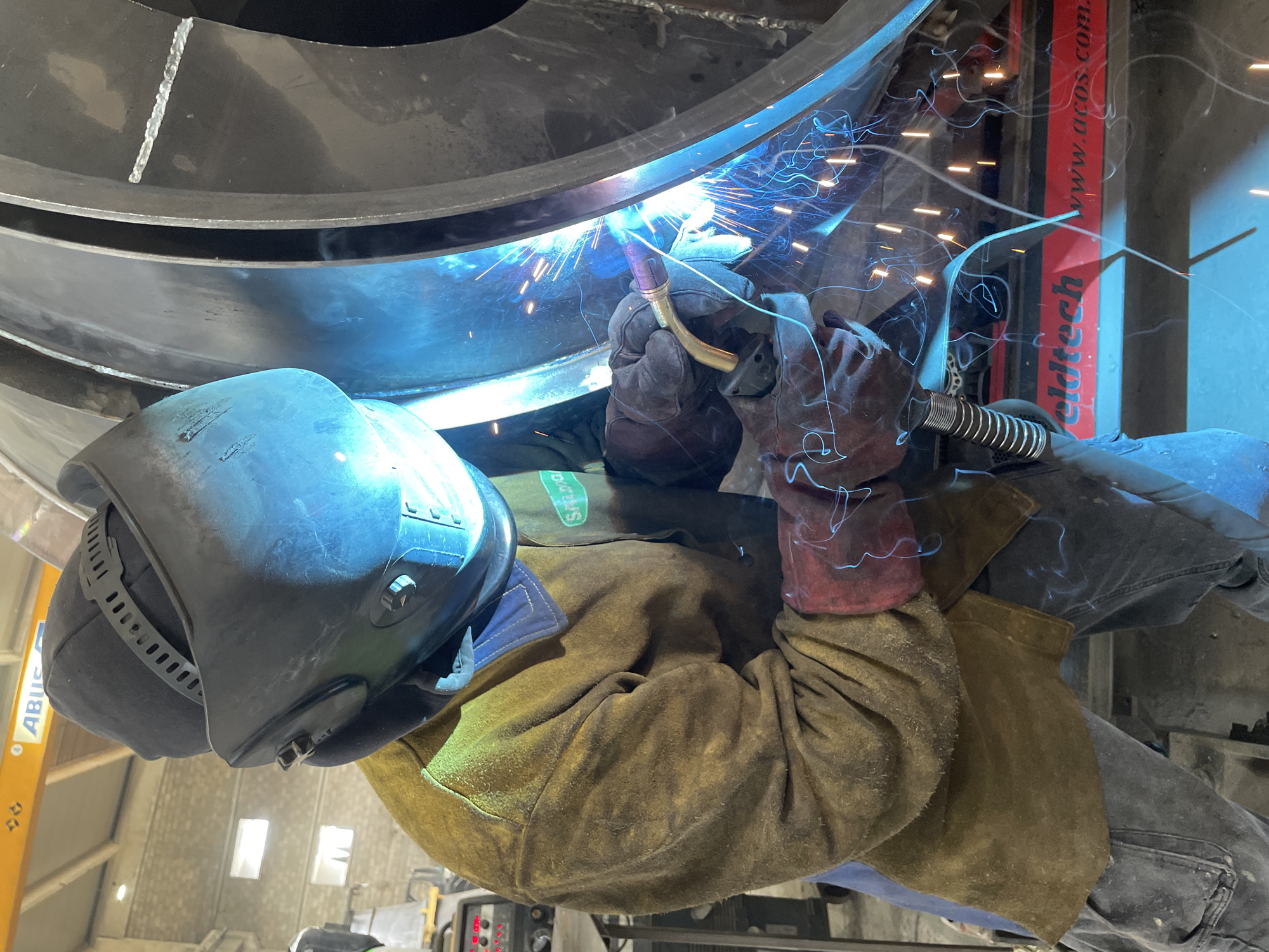 A welder in full protective gear is welding a large metal structure in an industrial setting. The welder is wearing a dark welding helmet, heavy gloves, and a tan jacket. Blue sparks and bright light emanate from the welding point. A welding machine with red and white signage can be seen in the background. The location is possibly inside a factory or workshop, identifiable by the construction materials and industrial equipment visible. Specific details indicating the location as Čeluga, Montenegro, are not discernible from the image provided.