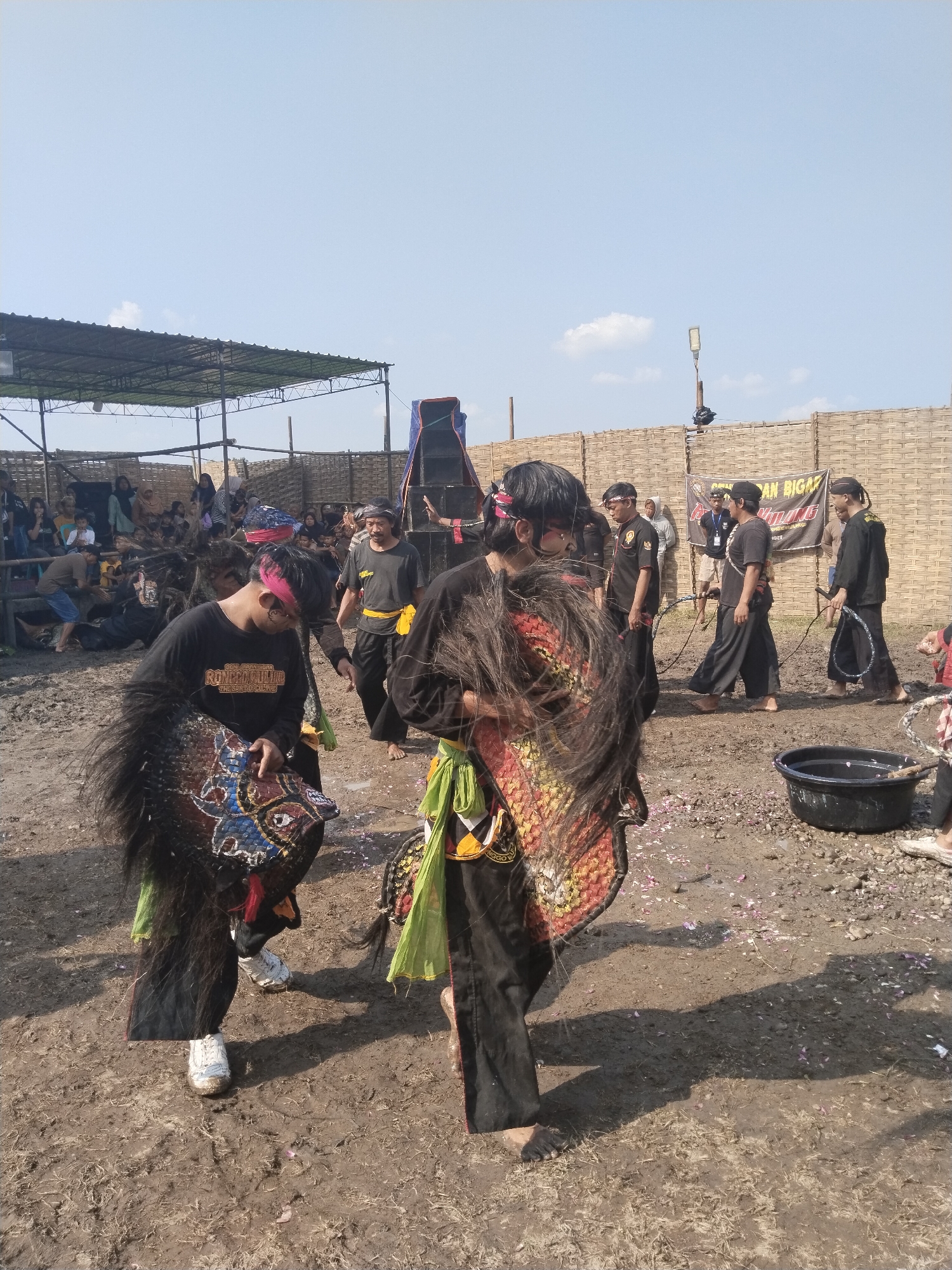 The image depicts a traditional cultural performance taking place in what appears to be a muddy open field in Kedawung, Indonesia. Two performers in black traditional attire, adorned with colorful fabrics and ornate masks, are the central focus. One performer is holding a mask with colorful patterns. They stand on muddy ground scattered with small, light-colored debris, possibly flower petals. Behind them, other people dressed in black uniforms are present, some holding what appear to be whips or rope-like objects. A group of spectators is seated under a makeshift shelter in the background. Beyond the immediate performance area, there's a woven barrier and a speaker system. A black tub filled with water is visible to the right. The environment suggests an outdoor cultural event in a rural or semi-rural setting.