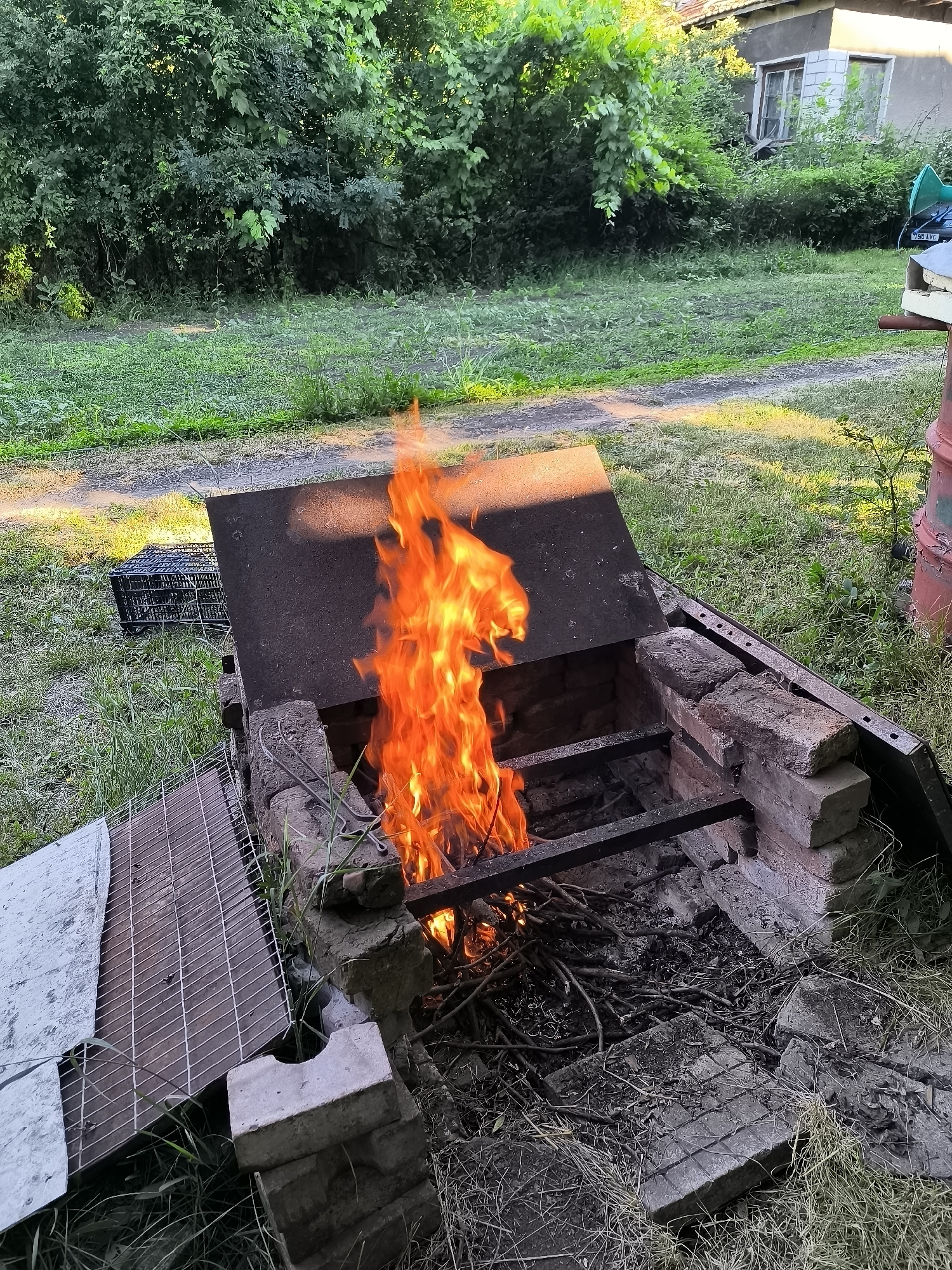 A mid-shot, eye-level, outdoor, slightly angled photograph captures a brick-built barbecue pit with a large, flat metal lid partially open, revealing a roaring fire within. The fire burns with vibrant orange and yellow flames, consuming a pile of dry sticks and debris at the bottom of the pit. The barbecue pit is constructed from weathered bricks, some of which are chipped and broken, suggesting age and use. Black metal grates are visible inside the pit, one resting on the side and another positioned above the flames. To the left of the barbecue, a metal mesh grill lies on the ground, next to a black plastic crate. In the background, lush green foliage forms a dense backdrop, with sunlight dappling through the leaves. Further back, a simple, somewhat dilapidated building with grey walls and windows is visible amidst the greenery. The ground surrounding the barbecue is covered in dry grass and scattered debris. The scene is set in a rural or semi-rural environment, likely a backyard or garden area. There is no indication of the specific location within the city of Sofia, Bulgaria, in the visual data provided. No people are present in the image.