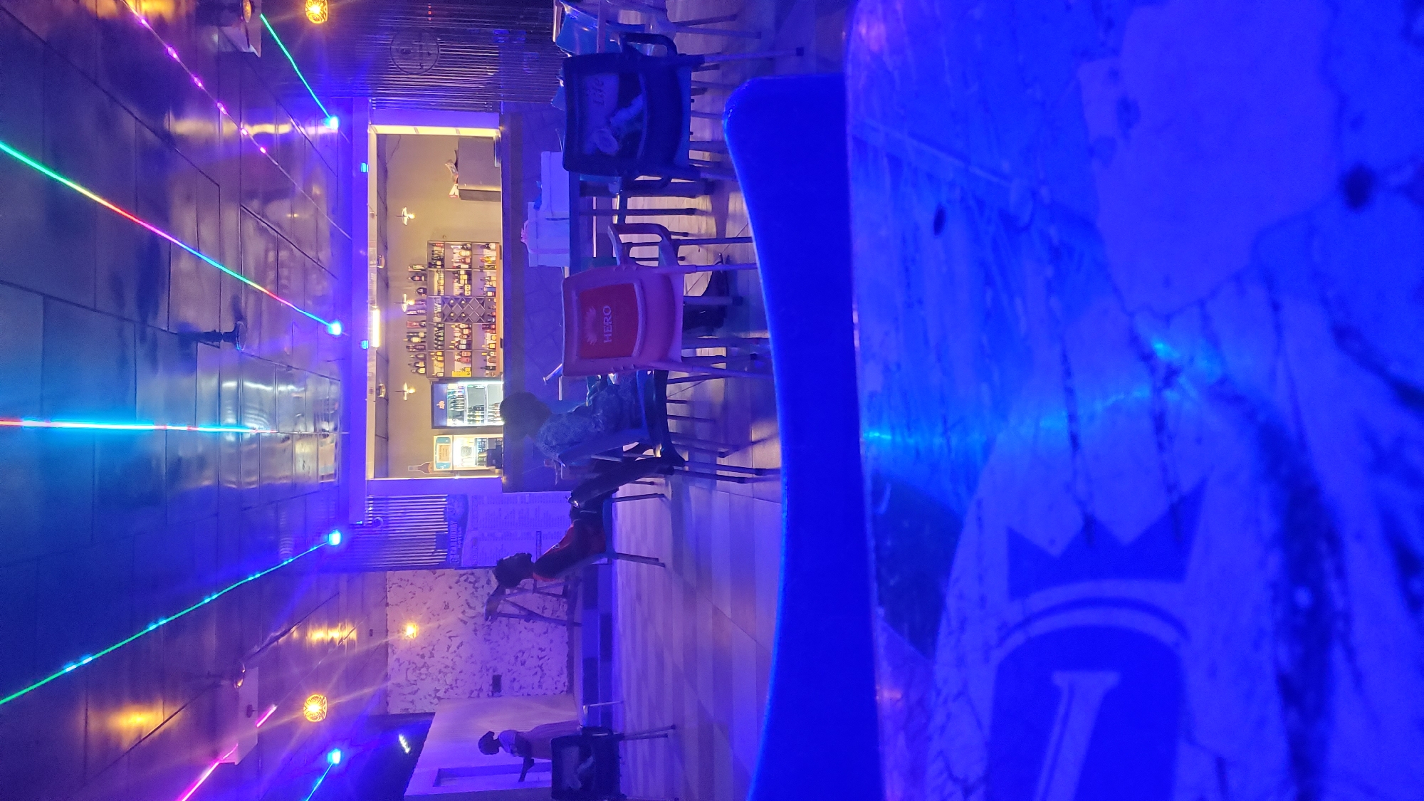 The image depicts the interior of a dimly lit establishment, possibly a bar or restaurant, in Akwanga, Nigeria. The scene is dominated by blue and purple lighting, with colorful neon strips running across the ceiling, casting an atmospheric glow. In the background, a well-stocked bar is visible, with shelves lined with bottles and a refrigerator. Several tables and chairs are arranged throughout the space.
In the mid-ground, there are several individuals. One person is seated at a table, leaning back in their chair. Another person is standing near a table, partially obscured. A third individual, a woman, is standing near a wall on the left side of the image, wearing a light-colored dress. The foreground is blurred, showing the surface of a table with a light blue tint, possibly indicating a protective covering. The overall ambiance suggests a lively, nighttime setting.