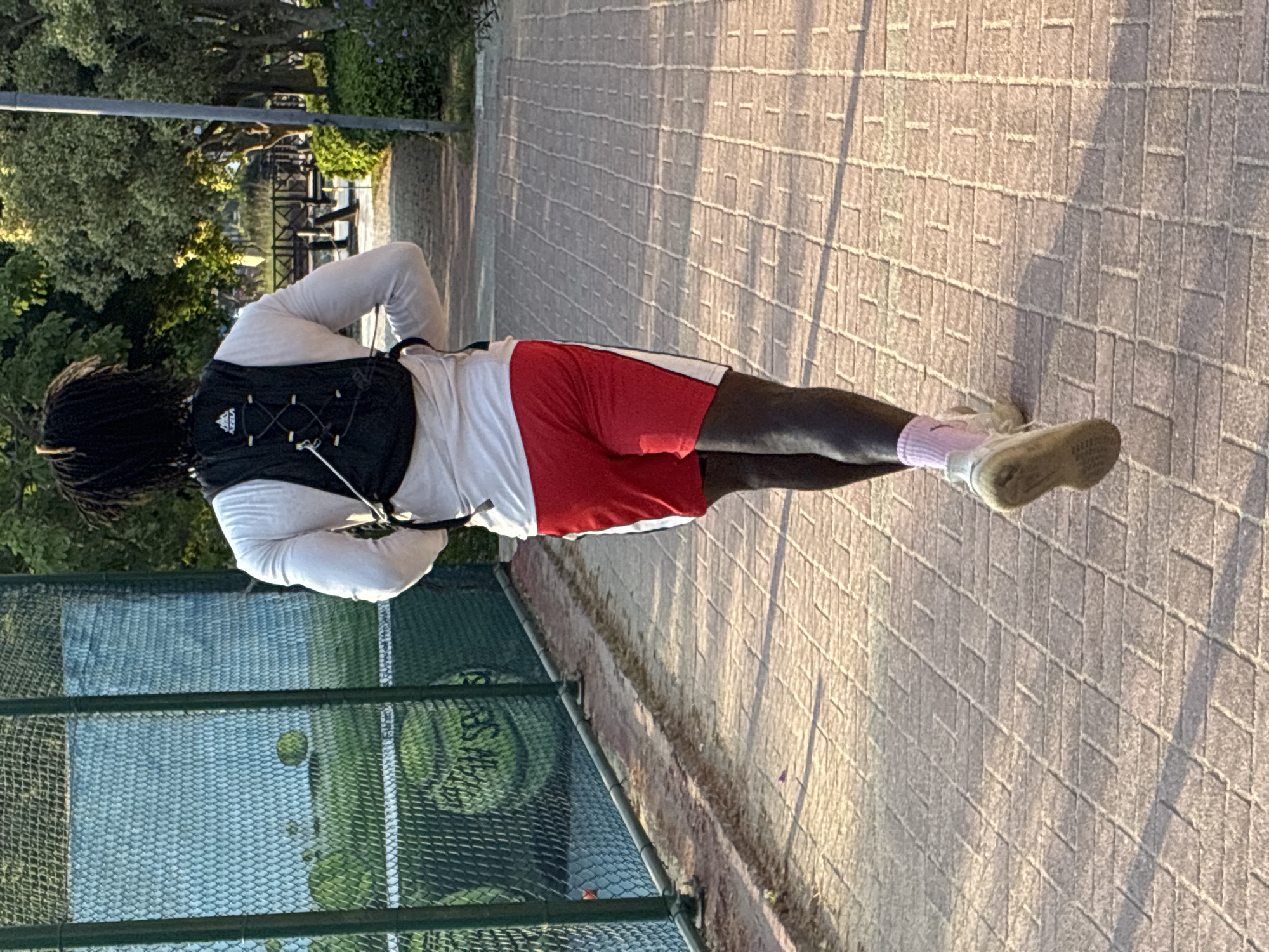 A Black male with dreadlocks runs away from the camera on a paved walkway. He is wearing a white long-sleeved shirt, a black backpack, red and white shorts, pink socks, and white athletic shoes. The walkway is lined with trees and bushes on the right and a tennis court with a green fence and large tennis balls on the left. In the background, there are more trees and a body of water with a wooden bridge. The scene appears to be outdoors during daylight.