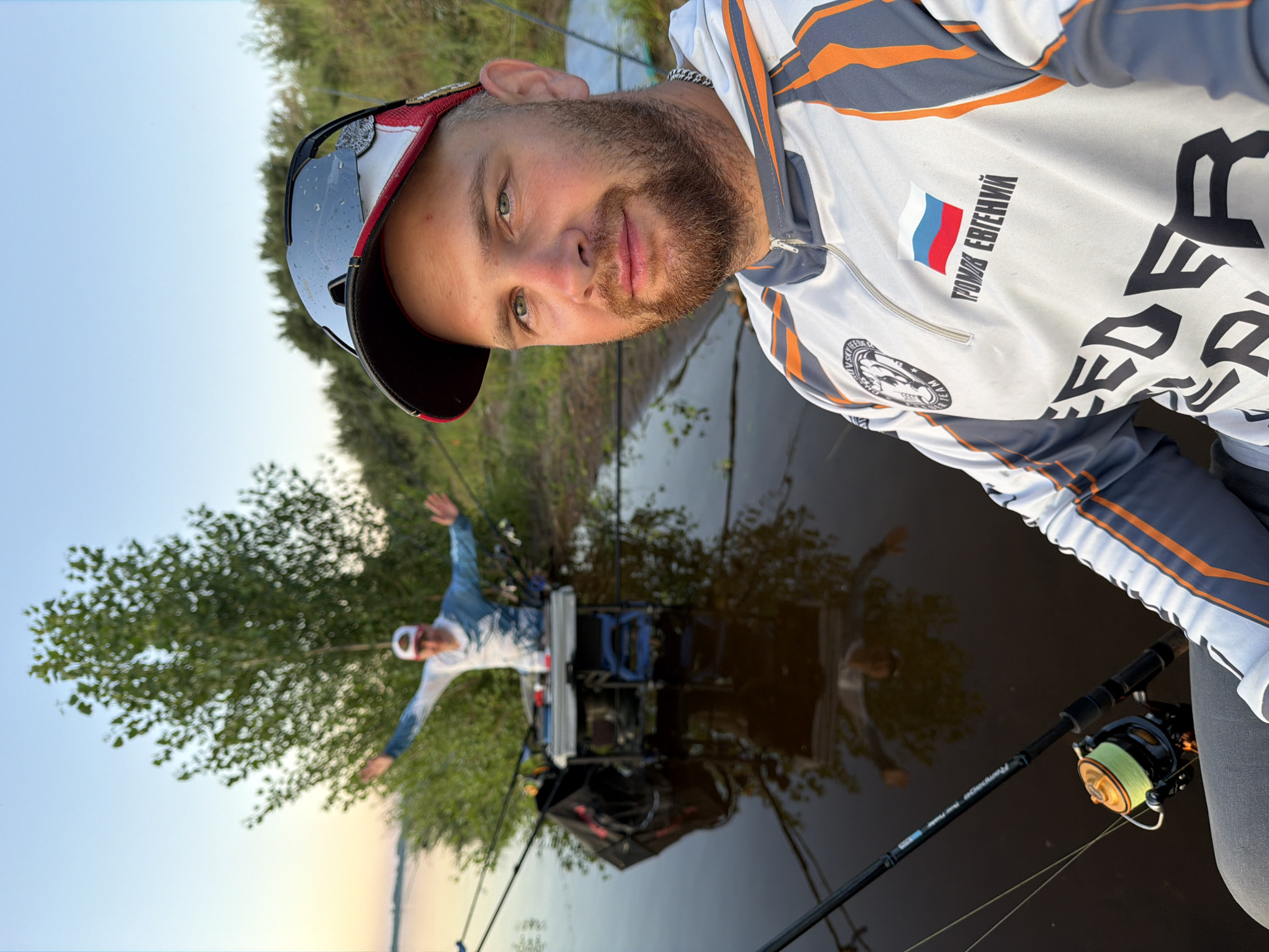 In the city of Tarasovo, Russia, two individuals are depicted near a body of water, likely engaged in fishing. The foreground is dominated by a man in a white, grey, and orange fishing jersey, wearing a baseball cap with sunglasses perched on top. He has a beard and is looking towards the camera. The jersey features Russian flag insignia and text. In the background, another person stands with arms outstretched, wearing a similar fishing outfit and a cap, facing away from the camera and towards the water. They are positioned behind a fishing station equipped with rods and tackle. The scene is set by a calm body of water reflecting the sky and surrounding trees and vegetation. The lighting suggests it is either early morning or late evening.