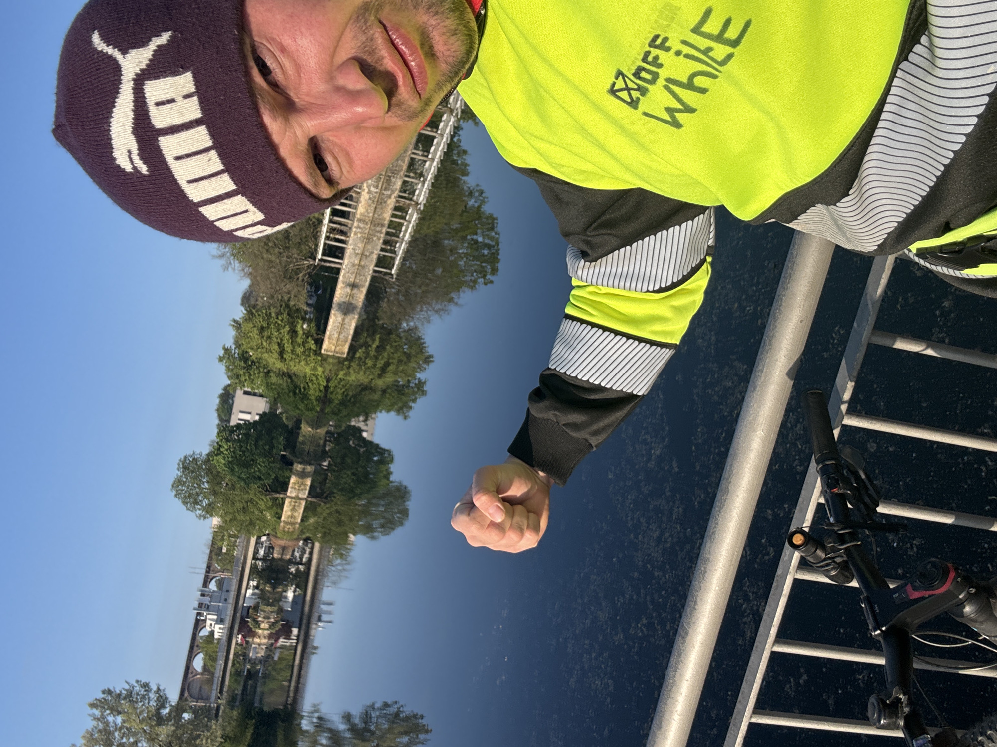 An outdoor selfie, presented rotated 90 degrees clockwise, captures a man against a scenic waterside backdrop in Bietigheim-Bissingen, Germany, on a clear, sunny day. The man, with a light beard and neutral expression, occupies the foreground. He wears a dark purple knit beanie with a white "PUMA" logo and a fluorescent yellow long-sleeved cycling top. The top features black sections, reflective white and grey striped patterns, and black text reading "OFF WHILE" with a small square logo on the chest. His right hand is clenched into a fist, extending forward.
A silver metal railing runs across the mid-ground, separating the man from a calm, dark blue body of water, likely a river or canal, reflecting the clear blue sky and distant scenery. On the far bank, lush green trees and various buildings are visible, including a prominent stone arch bridge or viaduct. The handlebars and front light of a bicycle are partially visible in the lower right, suggesting the man is on a bike ride. The bright light indicates it is daytime.