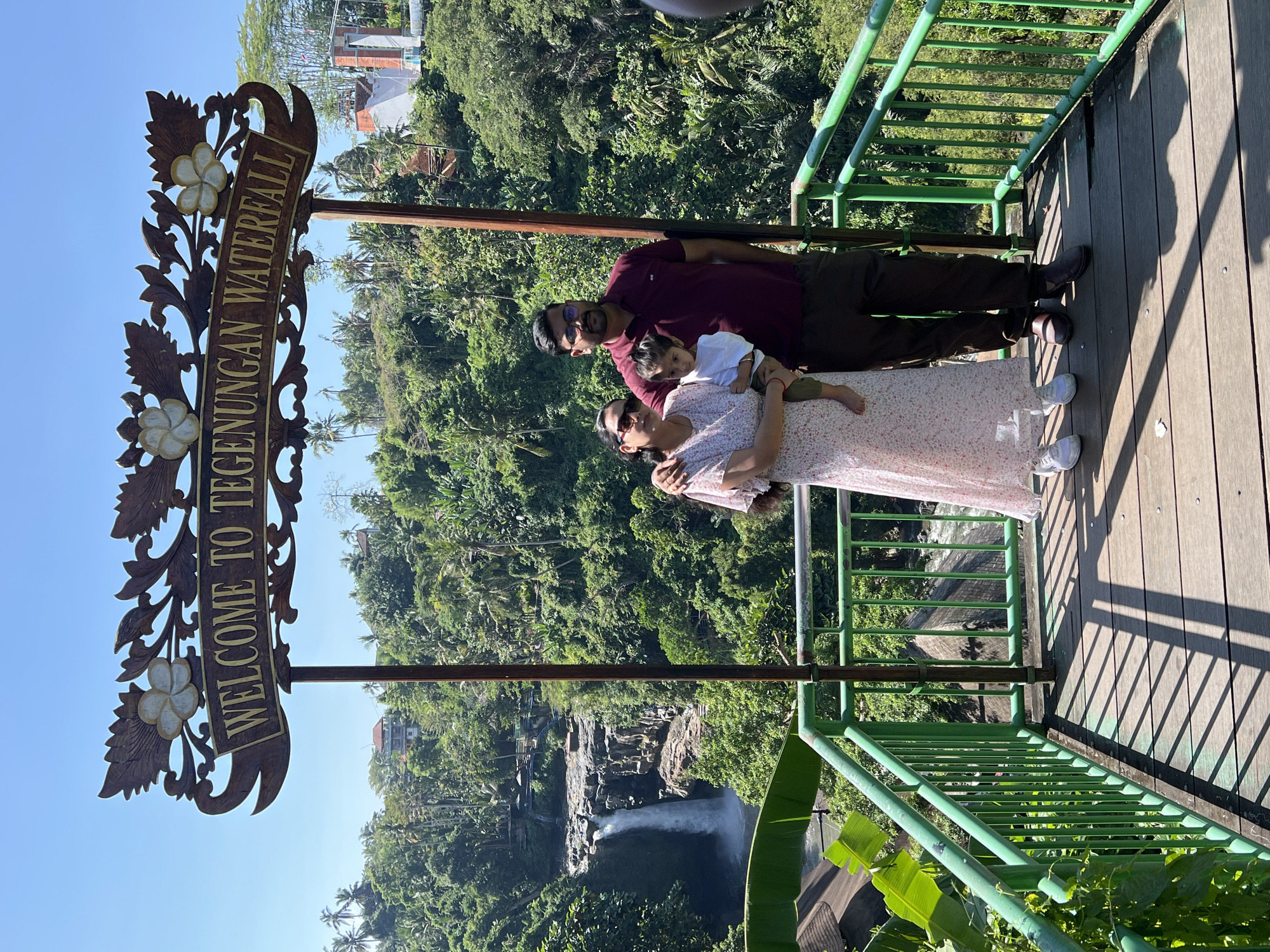 A family of three, two adults and a young child, stand on a wooden platform in front of a sign that reads "WELCOME TO TEGENUNGAN WATERFALL." The father, wearing a maroon polo shirt and dark pants, has his arm around the mother, who is in a floral-print dress and sunglasses. The child, dressed in white, is held by the mother. The platform is enclosed by a green metal railing, with shadows of the railing cast on the wooden planks. Behind the family, a lush green jungle slopes down to reveal the Tegenungan Waterfall, a tall cascade of water falling over rocky cliffs. The sky is clear blue. The scene is located at the Tegenungan Waterfall in Sukawati, Indonesia.