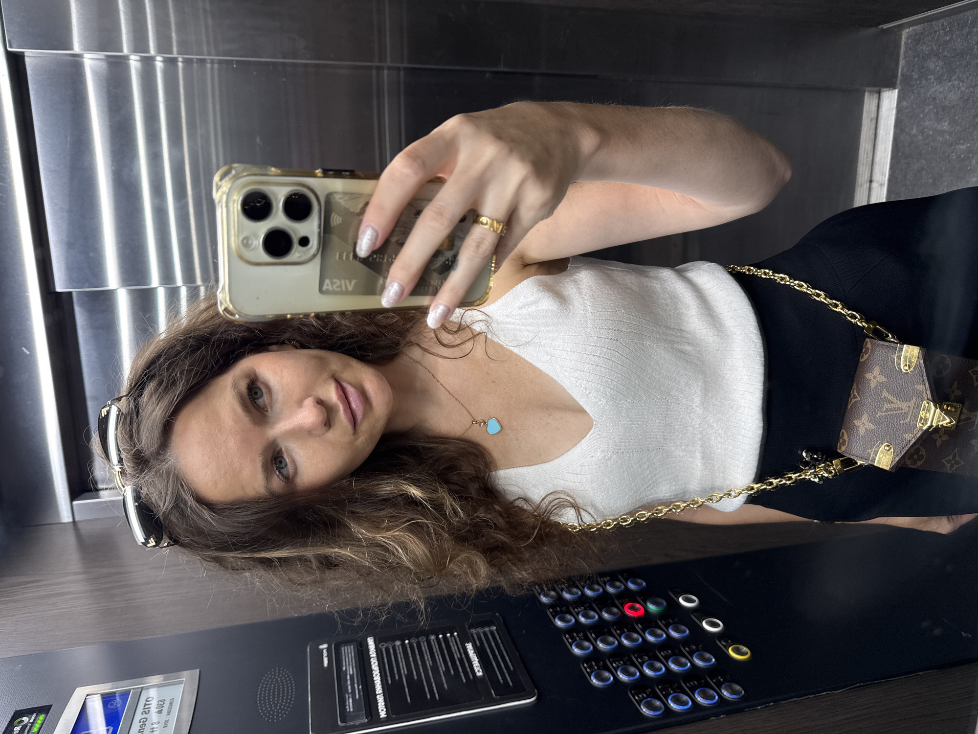 A woman with curly brown hair and sunglasses on her head takes a selfie in an elevator. She is wearing a white tank top, a black skirt, and a small Louis Vuitton purse with a gold chain strap. She has a light blue heart-shaped pendant necklace and a gold ring on her left hand. The elevator panel has numerous buttons with blue and red lights. The elevator walls are made of brushed metal. The image is a candid shot, likely taken in Moscow, Russia, given the elevator control panel which appears consistent with Russian standards.
