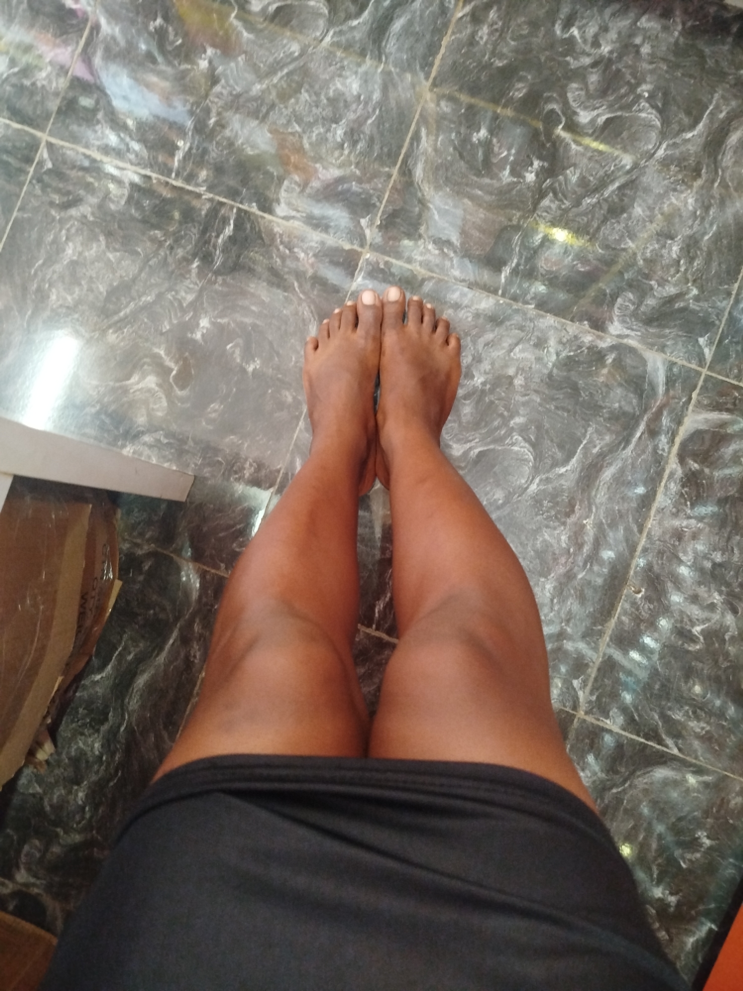 An overhead, slightly angled shot shows a person's legs from the knees down, standing on a tiled floor. The floor tiles are dark gray with prominent white veining, suggesting a marble or granite finish. The person is wearing a black skirt that ends mid-thigh. Their legs are bare and appear to be tanned. The feet are positioned together, with toenails visible. The left leg is slightly further forward than the right. To the left of the frame, partially visible, is the edge of a white piece of furniture and a brown cardboard box. The lighting in the scene appears to be artificial, with a bright reflection visible on the floor. The scene is indoors, likely within a residential or commercial space in Ugbowo, Nigeria.