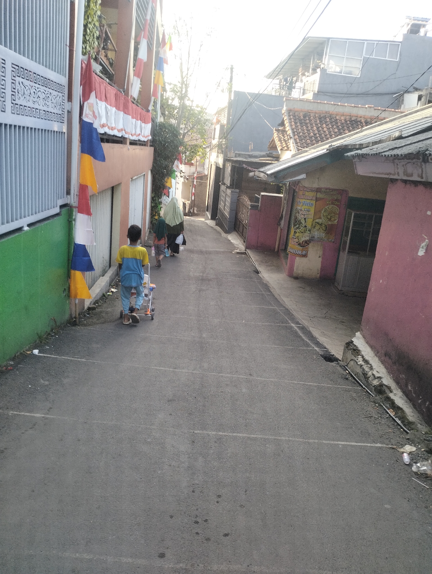 In the city of Kel. Pasir Jati, Indonesia, an outdoor scene captures a narrow, paved alleyway. Sunlight illuminates the scene, casting shadows. On the left, a green wall adjoins a white metal fence with a decorative pattern. Festive red, white, blue, and yellow flags are strung along a building's exterior on the left, cascading down towards the street.
A young boy, wearing a yellow and blue shirt and light-colored pants, walks away from the camera, pushing a small, multi-colored toy stroller. Behind him, two individuals, one wearing a light green headscarf and the other in darker clothing, are also walking away.
The alleyway itself is an asphalt surface with some visible white markings. To the right, buildings line the street. One building has a pink facade with yellow and white accents, featuring a sign with text and images that appear to advertise food. Another building to the far right displays a similarly pink exterior. The ground along the right side of the alley shows a concrete curb with some debris, including plastic bottles and trash. Overhead, electrical wires are visible. The environment suggests a residential or mixed-use urban setting.