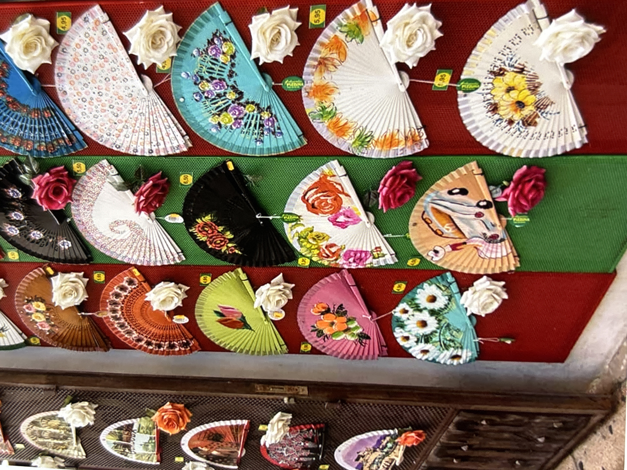 The image depicts a vibrant display of decorative fans, likely for sale, on the exterior of a shop in Orihuela, Spain. The fans are fanned open and arranged in rows on red and green fabric backdrops, creating a colorful and eye-catching presentation.
On the left side of the image, a dark wooden door with ornate metalwork stands partially ajar, revealing a textured surface behind it. Several fans are attached to this door, featuring designs that include painted architectural scenes, floral motifs, and what appear to be portraits. White and orange fabric roses are affixed to some of these fans.
To the right of the door, a large collection of fans is displayed on vertically oriented red and green fabric panels. These fans exhibit a wide range of designs, from intricate floral patterns in various colors, to graphic representations of insects and more abstract designs. Many of the fans have small, square price tags attached, indicating they are merchandise. White fabric roses are also used as embellishments on some of these fans. The overall impression is that of a lively market stall or a shopfront showcasing traditional Spanish crafts. The ambient lighting suggests it is daytime, likely outdoors.