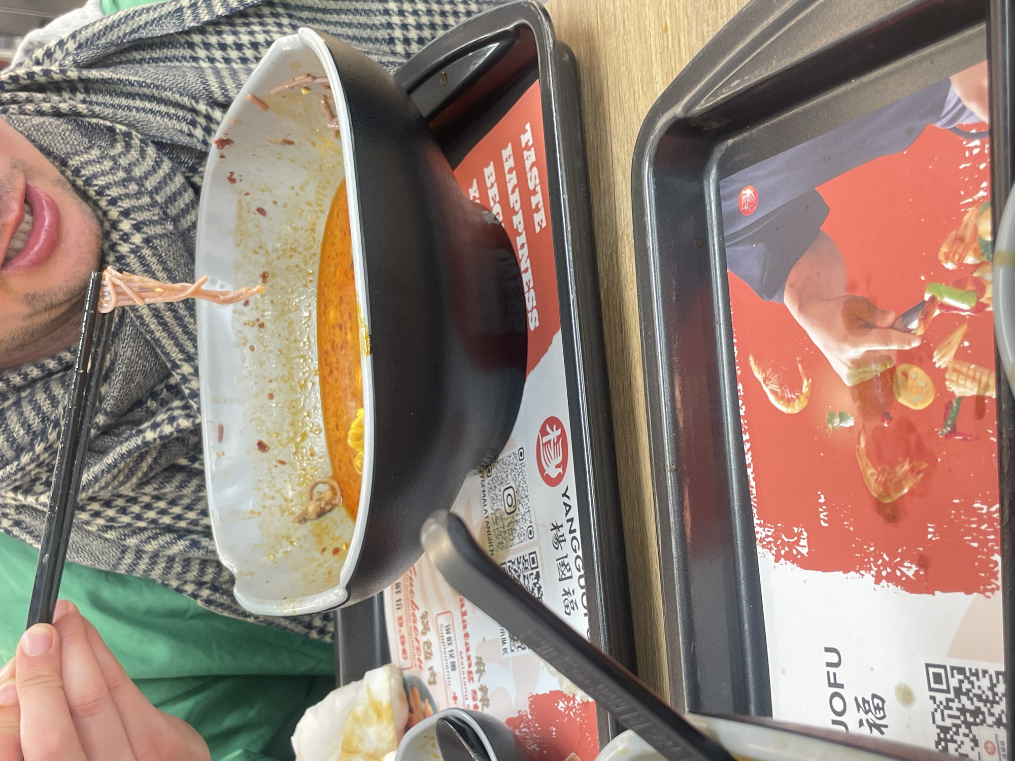 The image shows a person eating at a Yangguofu Malatang restaurant. The person is holding chopsticks with food in them, and there is a bowl of food on a tray in front of them. The tray has the Yangguofu logo and a QR code on it. Another tray with an image of someone cooking is partially visible. The location is not visible in the image, and there is no information to identify if the restaurant is located in Barcelona, Spain.