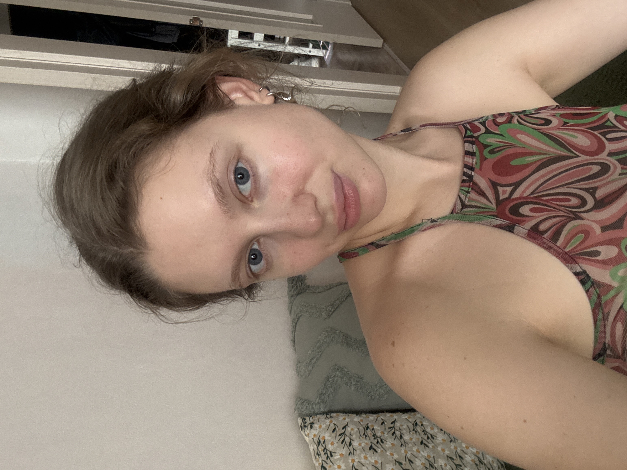 A woman with light skin and blue eyes is shown in a close-up, bust shot, facing the camera. She has light brown hair pulled back and is wearing a patterned halter top with swirling designs of pink, green, and brown. Her left ear has a silver hoop earring. She is leaning against a couch with two patterned pillows behind her; one pillow has a floral print, and the other is a textured green pillow. The background shows a doorway with a white door and a glimpse of a closet or room beyond, with the floor appearing to be wood. The setting is indoors, likely a residential environment, with neutral-colored walls. There is no specific information about the location being in Izhevsk, Russia.