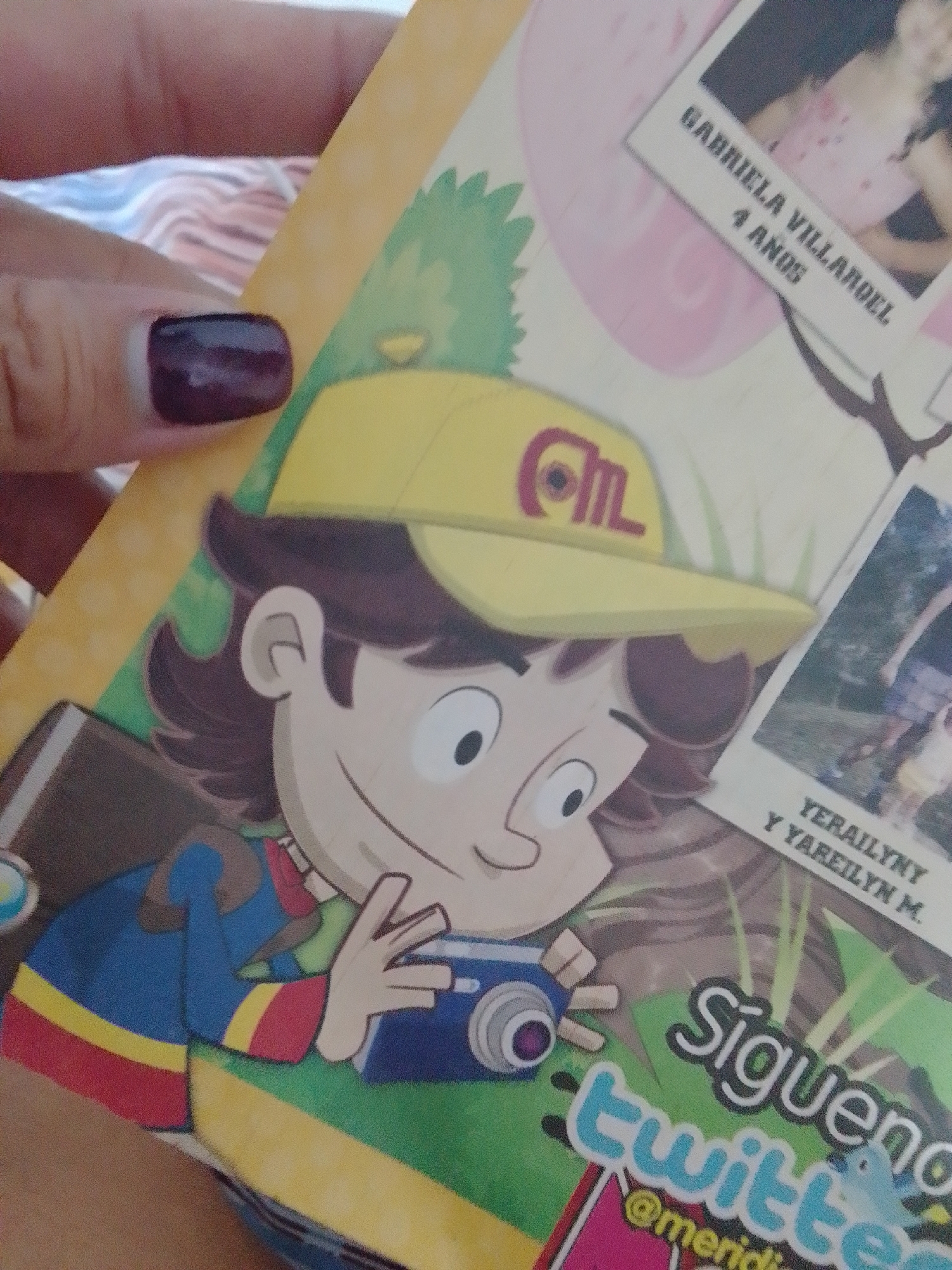The image is a close-up of a person holding a children's book or magazine. The book features a cartoon illustration of a young boy wearing a yellow cap with a red emblem and a blue and red striped shirt. The boy is holding a blue and silver camera. Text on the book includes "GABRIELA VILLARDEL 4 ANOS", "YERAILINY Y YAREILYN M.", and "Siguenos twitter @meridi". The background of the book has green and brown abstract shapes suggesting nature. A person's thumb with dark purple nail polish is visible at the top left, holding the book. The lighting is slightly diffused. There is no visible indication or context relating to the city of La Tagua, Venezuela within the image itself.