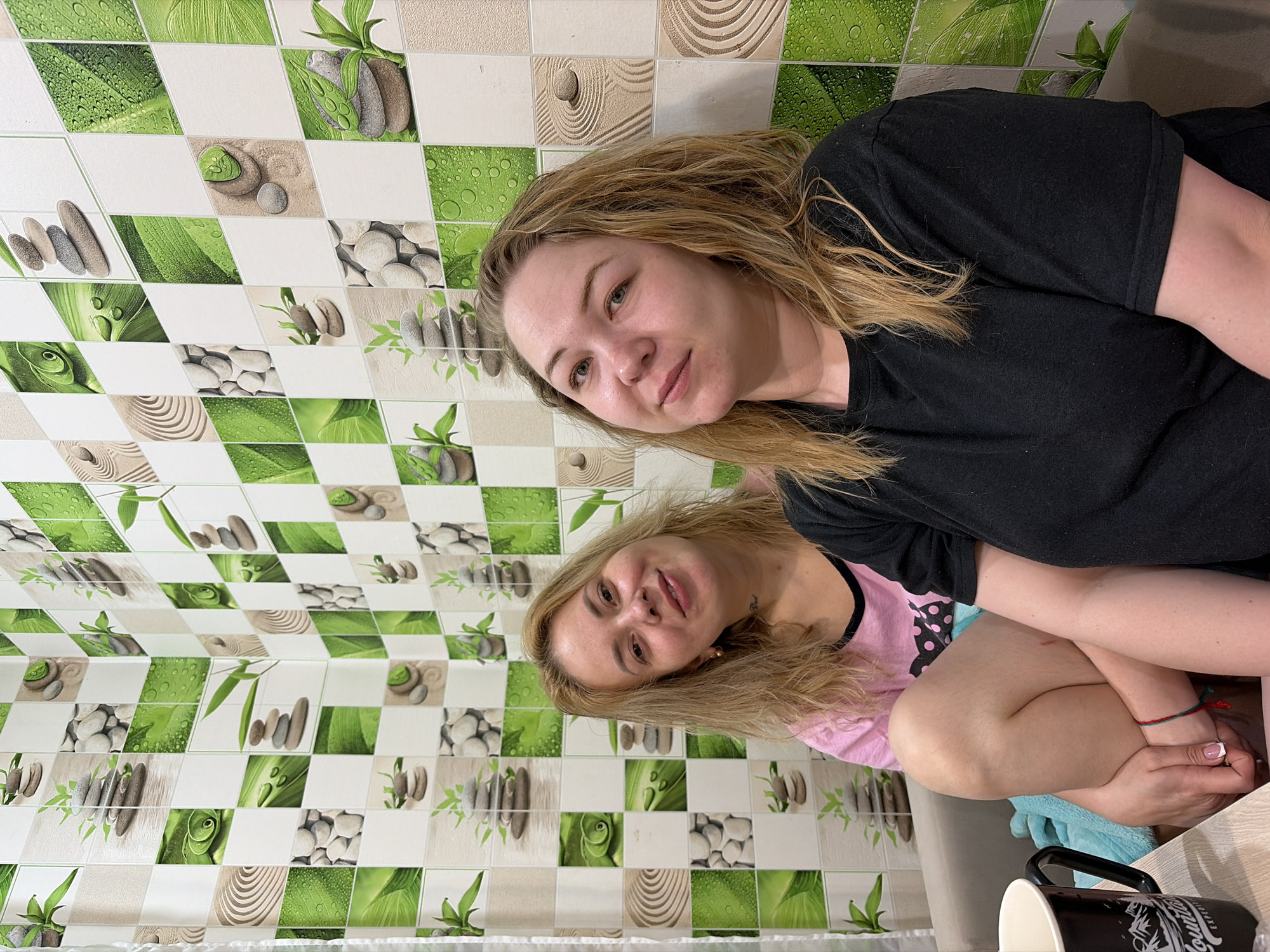 Two women are positioned in front of a wall covered with square tiles featuring green and white patterns of leaves, stones, and sand. The first woman wears a black t-shirt and has blonde hair. She is placed in the upper right part of the image, and her face is visible. The second woman is seated below the first, wearing a pink t-shirt with black details. She has blonde hair. A black mug sits on a table in the lower right corner of the frame. The location is Noviy, Russia.