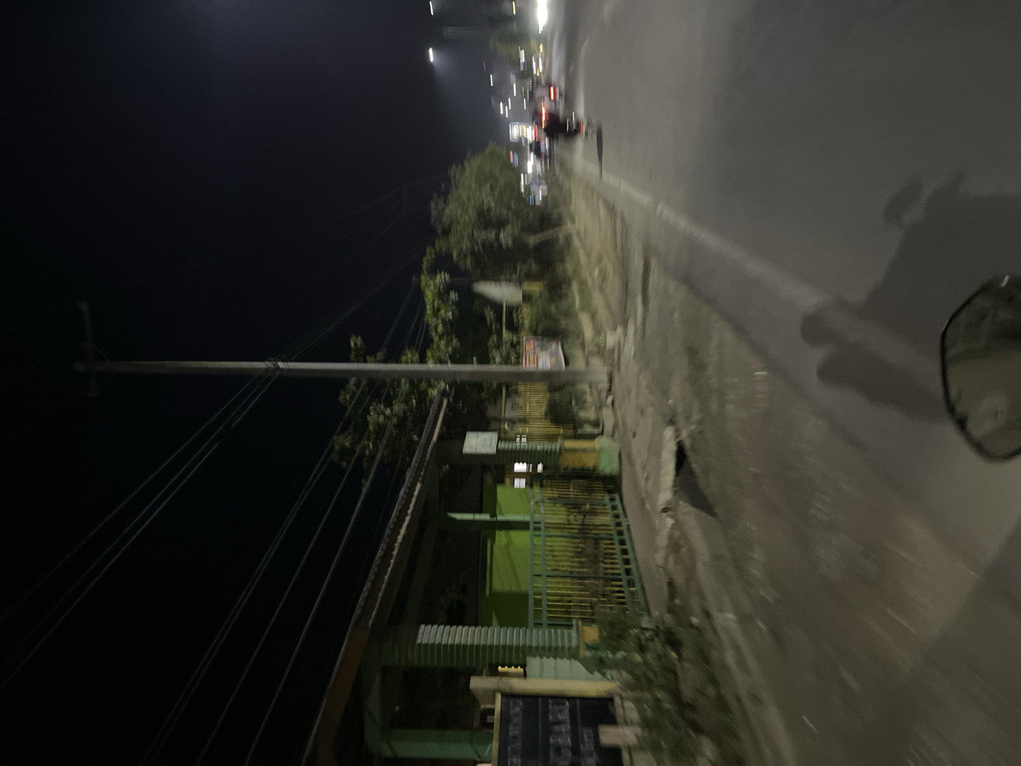 The image depicts a street scene at night in Tanjungkarang, Indonesia. The perspective is from a moving vehicle, likely a motorcycle, as evidenced by the presence of a side mirror in the lower right corner. The road is visible on the right side, appearing wet and reflecting the lights from vehicles and streetlights. On the left side of the image, there is a utility pole with numerous wires extending across the frame. Beyond the pole, a building with green fencing and what appears to be a dark-colored roof is visible. The building has multiple windows, some of which are illuminated. Further down the street, other vehicles and buildings are blurred due to motion and the low-light conditions. The overall impression is of a typical urban night scene, with the activity of traffic and the presence of infrastructure like utility poles and buildings. The time of day is clearly night, and the wet road suggests recent or ongoing rain. There is no discernible emotion conveyed, and any visible text is illegible due to blur and distance.