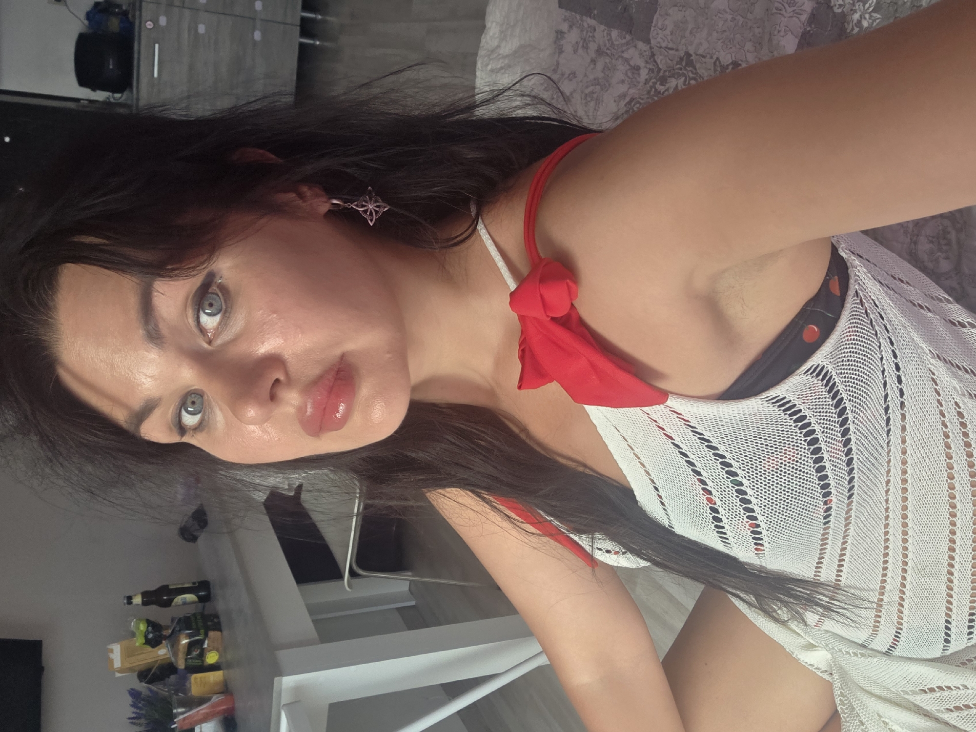 The image shows a woman with dark hair and blue eyes. She is wearing a white knitted cover-up over a black and red polka dot bikini top. The cover-up is tied with a red bow on her shoulder. She is also wearing a silver earring with a pink stone. The background shows a blurry interior scene with a table, chairs, and cabinets. There are no specific elements in the image that suggest the location is Sochi, Russia.