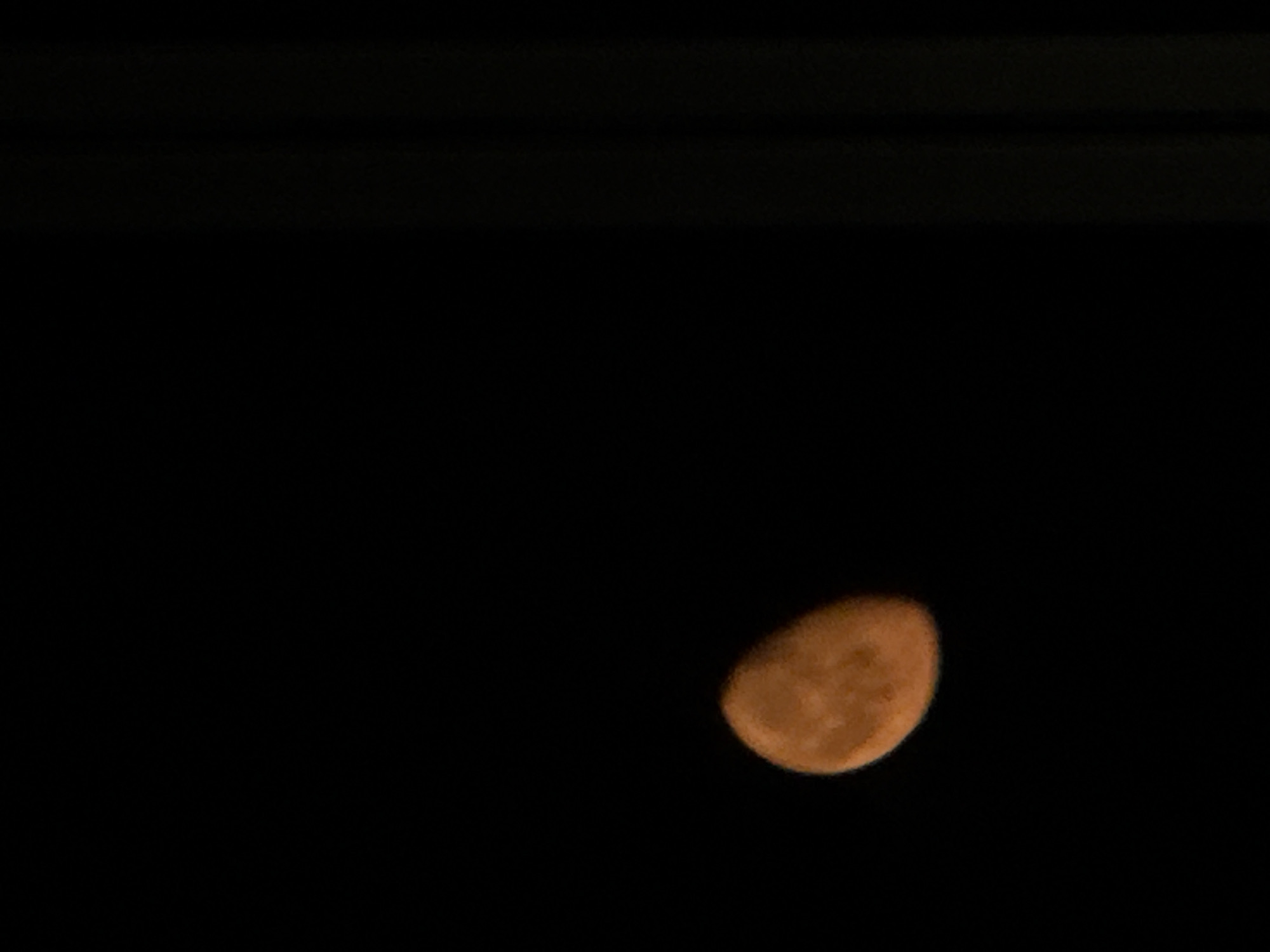 A partially illuminated moon, appearing orange in color, is visible in a dark sky. The moon is positioned in the lower left quadrant of the image. To the right of the moon, there are faint vertical lines, which appear to be part of a structure or building. The background is uniformly black, indicating a nighttime scene. The image does not contain any discernible features that would allow for the identification of the city of Tbilisi, Georgia.