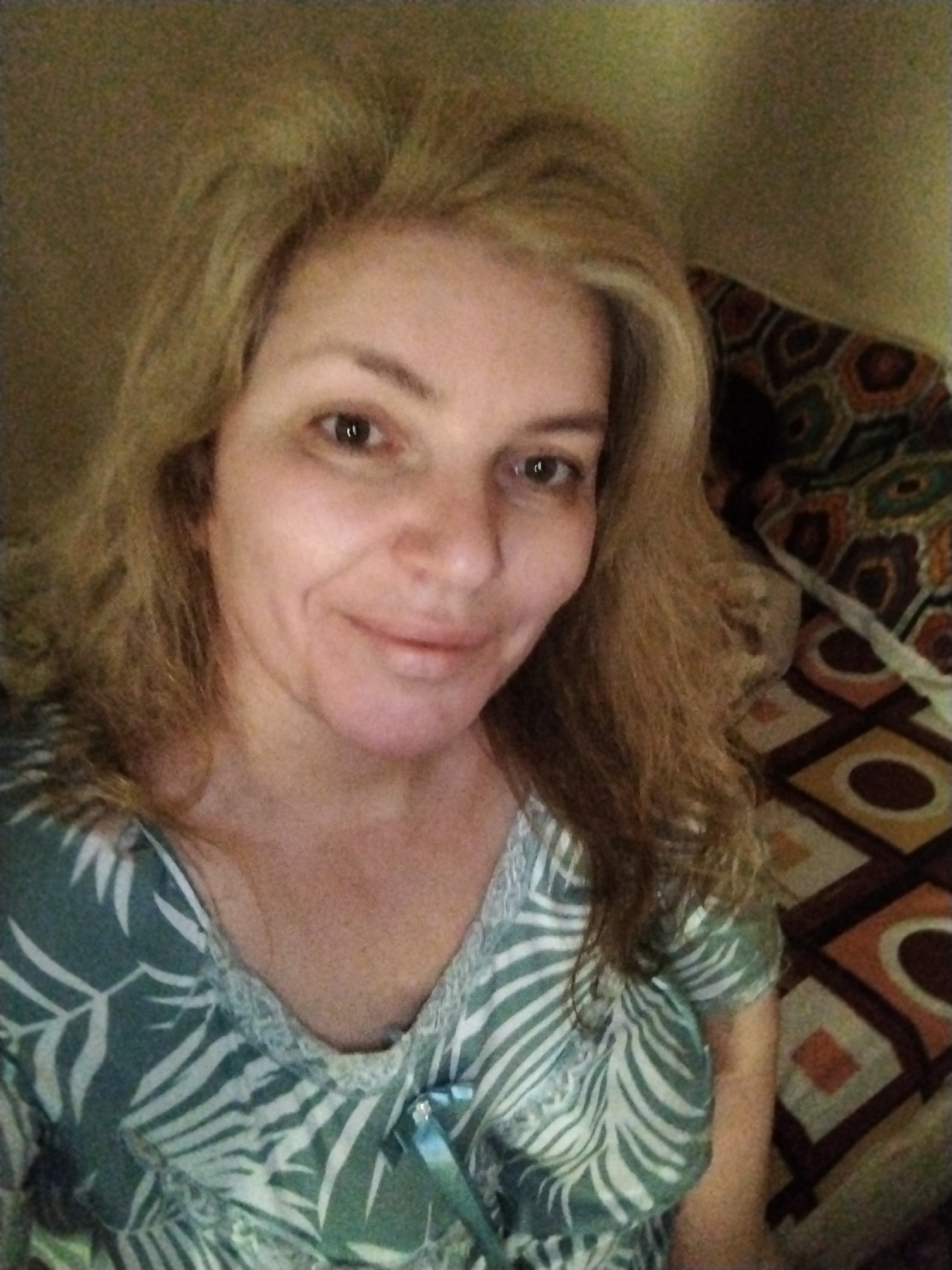A middle-aged Caucasian woman is centered in the frame, facing the camera. She has shoulder-length, blonde hair that appears somewhat tousled. Her facial features are visible, and she has a faint smile.
She wears a short-sleeved top with a light green background and white leaf and zebra-like stripes. The neckline has a lace trim and a small blue ribbon detail.
Behind her, a bedspread with a geometric pattern of squares and circles in shades of brown, white, and tan is visible. The setting seems to be indoors. The lighting is somewhat diffused, and the image has a slightly grainy quality, suggesting it may have been taken in lower light or with a basic camera. The location in the city of Al Mas'oudi, United Arab Emirates is impossible to determine from the image.