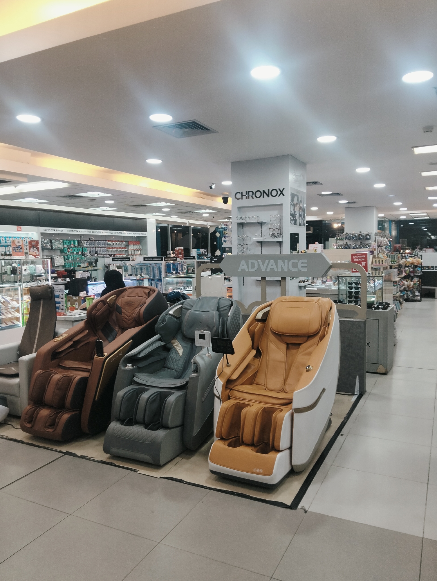 In the City of Medan, Indonesia, an interior scene depicts a retail environment, likely a department store or electronics store, with rows of massage chairs prominently displayed in the foreground. Three massage chairs are visible: a brown one on the left, a grey one in the middle, and a tan and white one on the right. In the background, shelving units filled with various products, including electronics and accessories, are arranged. A sign for "CHRONOX" is visible on a pillar. The lighting suggests an indoor, possibly artificial, illumination. The floor is tiled. A person wearing a black head covering is partially visible behind the brown massage chair.