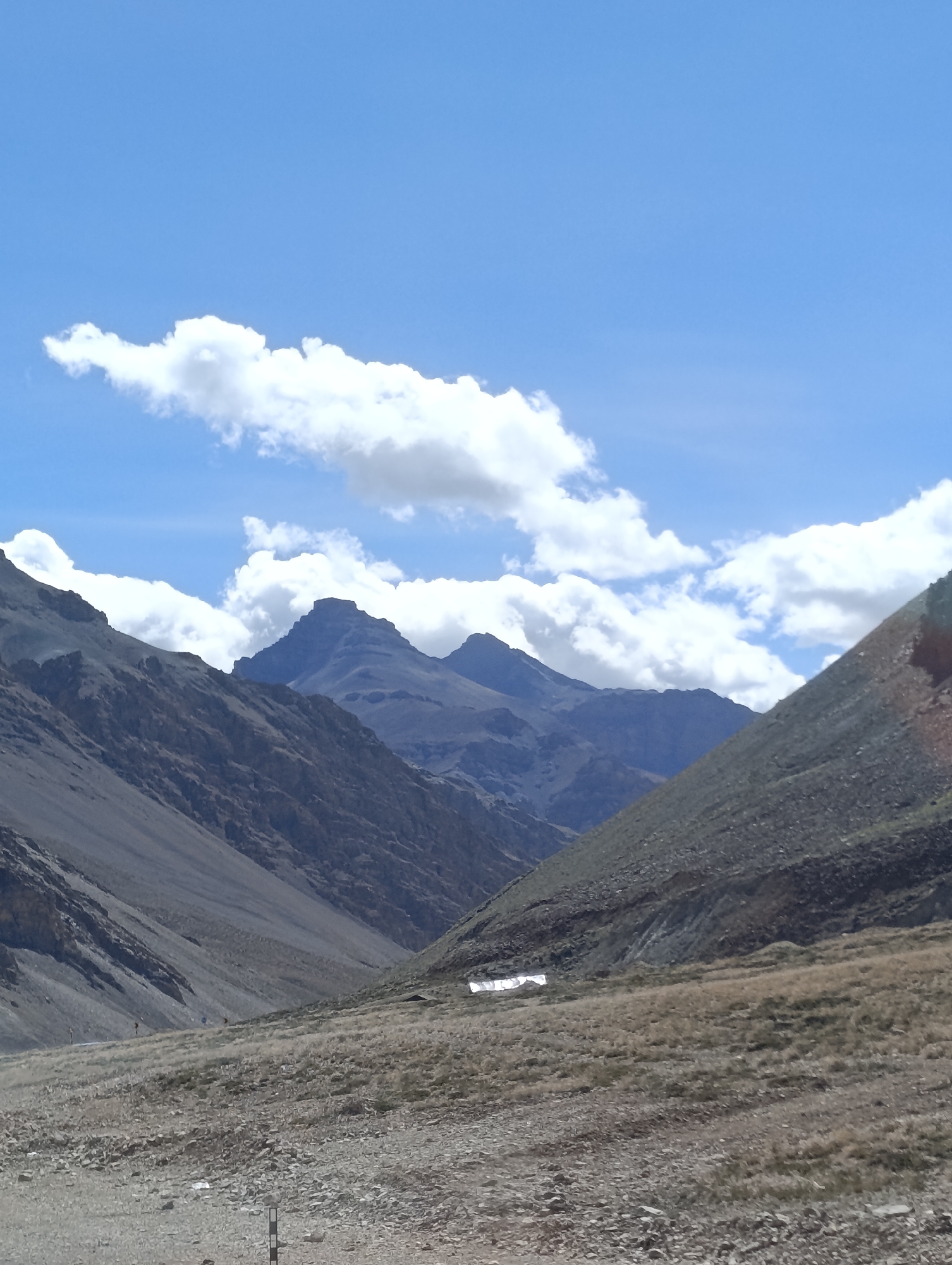 A wide, vertical shot captures a mountainous landscape under a bright blue sky with scattered white clouds. The foreground is a gravelly, uneven terrain with sparse, dry grass. In the middle ground, a valley opens up, flanked by steep, rocky mountainsides. A small, white structure, possibly a building or shelter, is visible in the valley. Further in the distance, more layered mountains rise, their peaks sharp against the sky. The overall impression is one of a remote, arid, and high-altitude environment. The location is identified as Dhar Sajam, India.