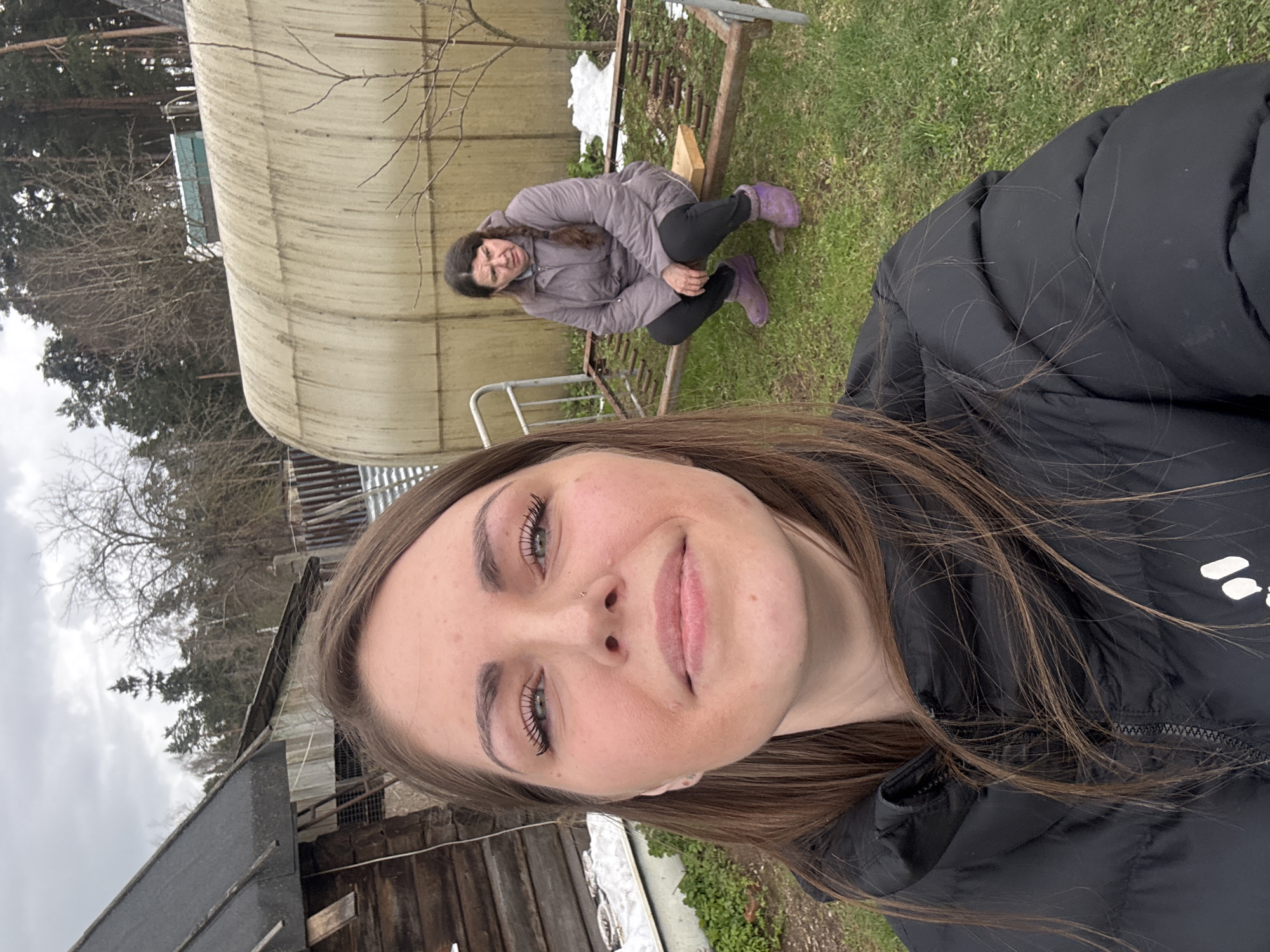 The image is a selfie taken outdoors. Two women are visible. The woman taking the selfie is in the foreground, wearing a black puffer jacket. Her hair is brown. In the background, another woman is crouching near a structure resembling a greenhouse, wearing a grey jacket and purple boots. There is snow on the ground in places. The environment includes trees, possibly at the edge of a forested area, and wooden structures. The location is Razdol'ye, Russia.