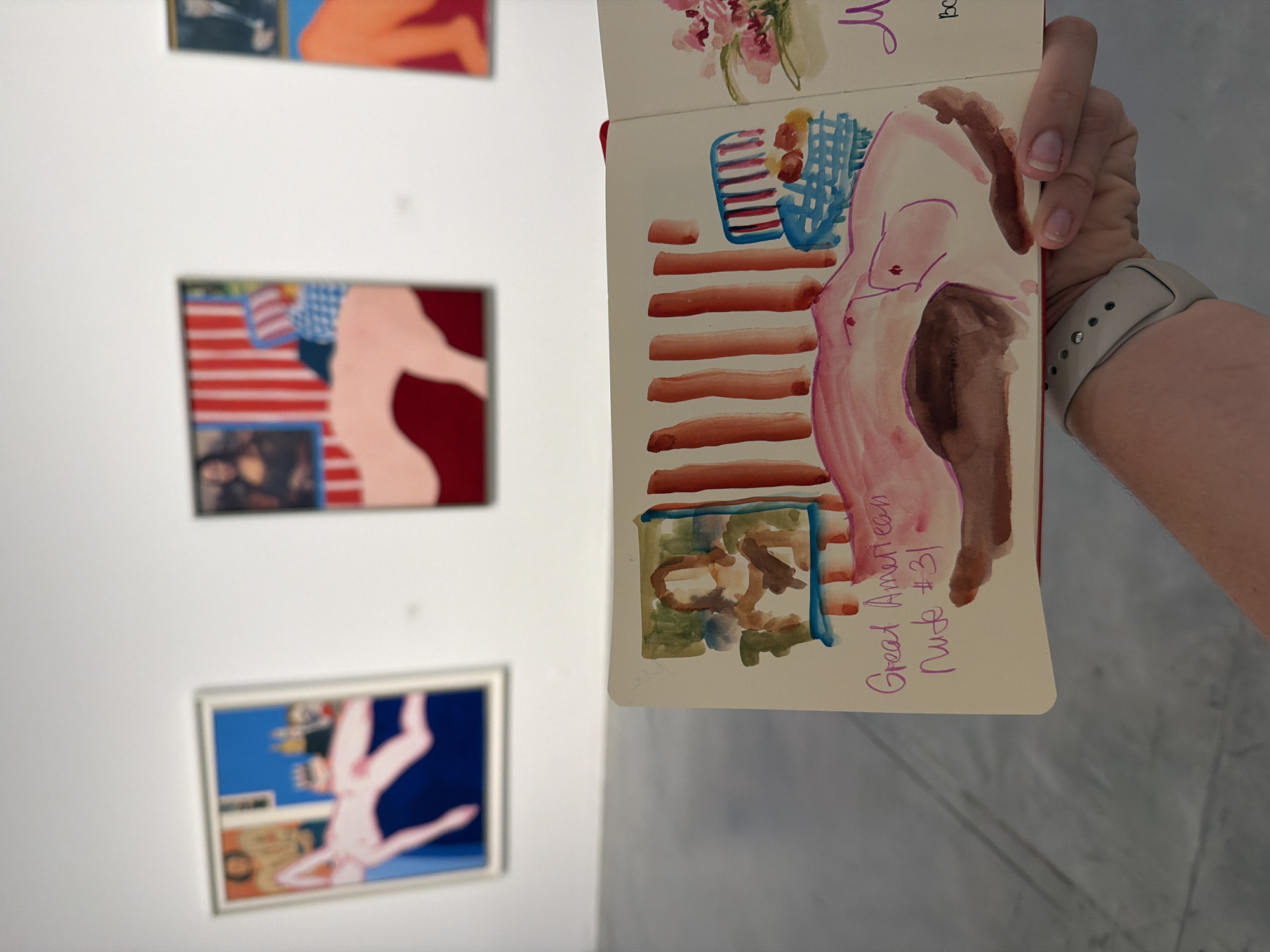 The image shows a person holding an open sketchbook at an art exhibition. In the foreground, the sketchbook displays a watercolor sketch of a reclining nude figure, with the text "Great American Nude #31" written below it. In the background, three large framed artworks are visible on a white wall. These artworks appear to be paintings, with one depicting a pink reclining figure against a blue background, another featuring red and white stripes and a Mona Lisa-like portrait, and a third showing an orange figure. The overall setting suggests an indoor exhibition space, likely a gallery. There is no information in the image to indicate the location is Tel-Aviv, Israel.