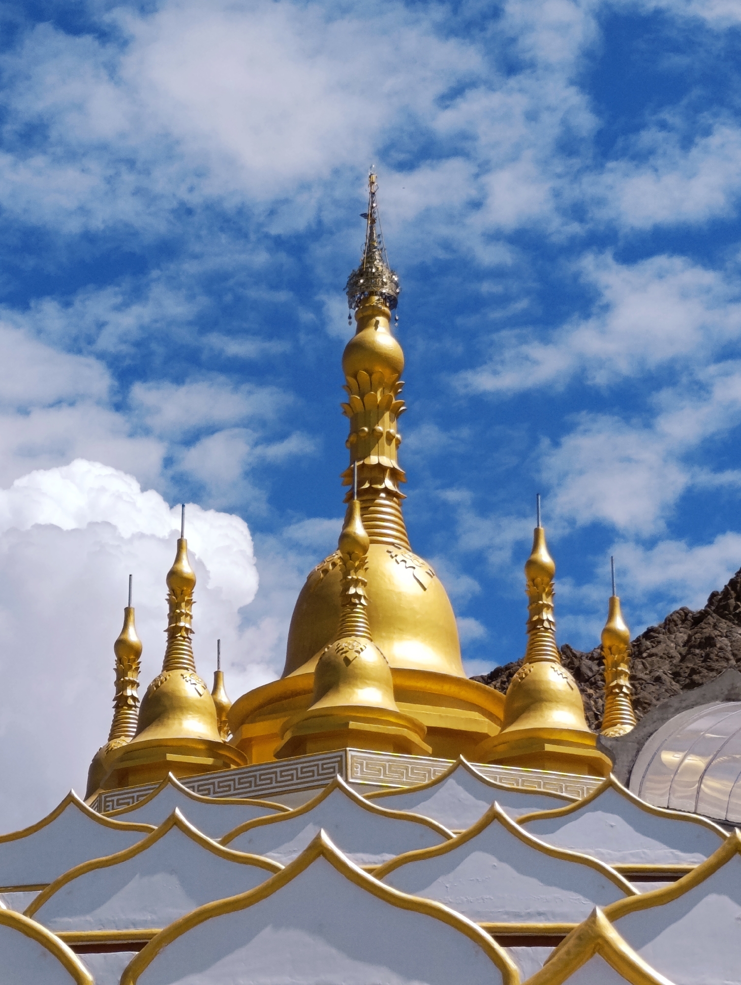 The image shows the golden stupas of the Shey Monastery, located in Shey, India. The monastery is situated in a mountainous region with a clear blue sky dotted with white clouds. The stupas, which are Buddhist reliquary monuments, are the main subject of the image, with their gold surfaces reflecting the sunlight. The foreground features white and gold decorative elements of the monastery's architecture. The rocky mountains are visible in the background on the right side of the image. No people are visible in the photograph.