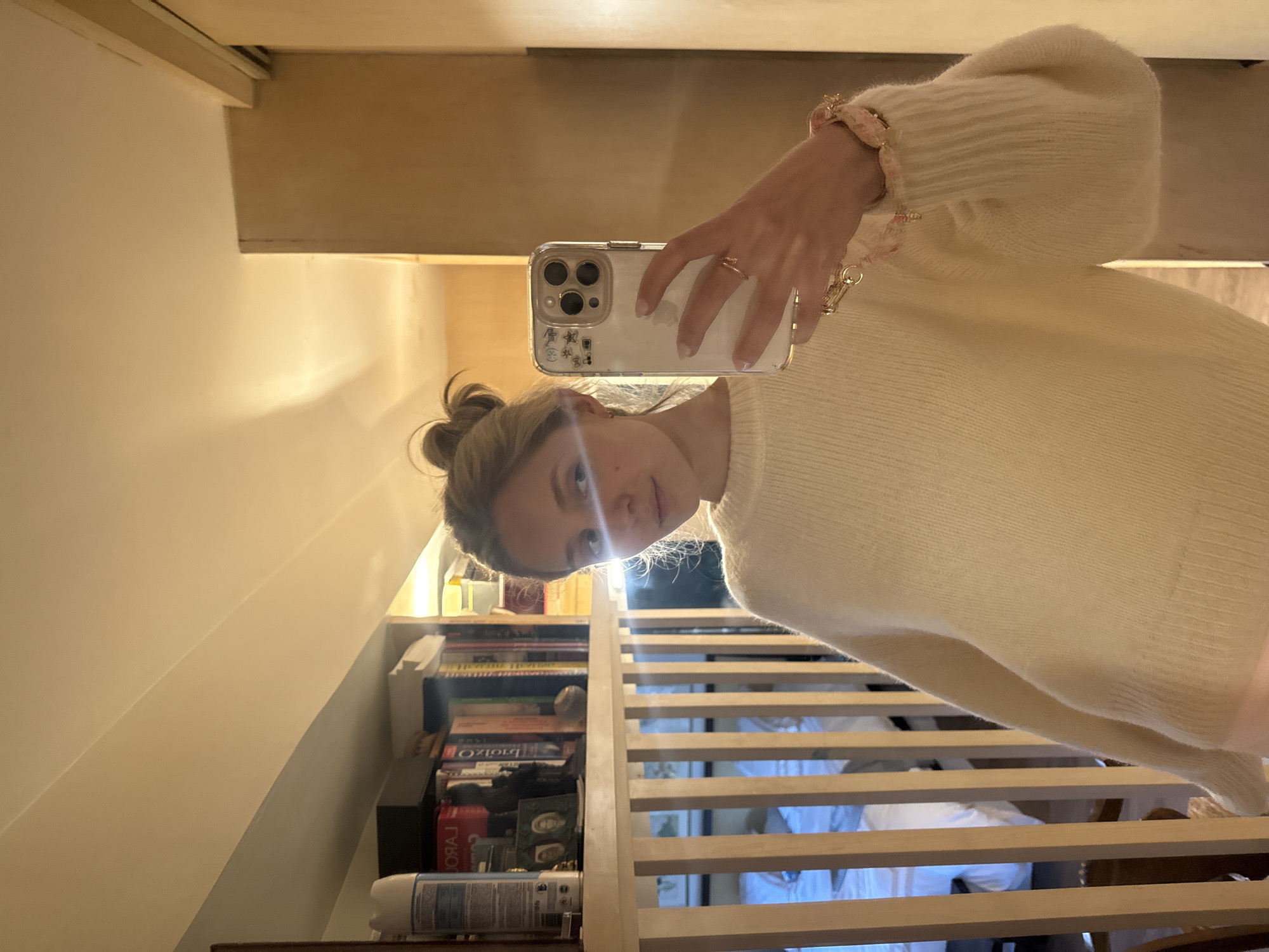 The image shows a young woman taking a mirror selfie in a room. She has blonde hair styled in a messy bun and is wearing a cream-colored sweater and pink pants. She is holding a transparent phone case with stickers on it and is wearing rings and a bracelet on her right hand. In the background, there are bookshelves filled with books and a glimpse of a bed through a wooden railing. The lighting suggests it is indoors, possibly in a home environment. There is no indication of the city of Ciudad de México, Mexico.