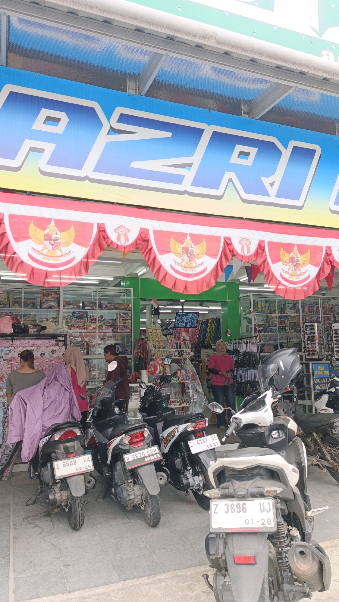 The image shows the exterior of a shop in Desa Karangjaladri, Indonesia. A large blue banner with the word "AZRI" is displayed above the entrance. Red and white pennants with the Garuda Pancasila symbol hang below the banner. Several motorcycles are parked in front of the shop. Inside the shop, shelves are stocked with various items, and a few people are visible. The overall scene depicts a commercial establishment in a bustling area.