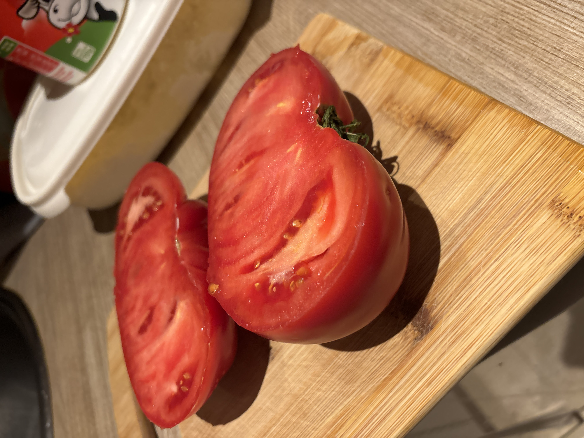 A halved, ripe, heirloom tomato lies on a bamboo cutting board next to a cut slice. To the left is a white plastic container of cream cheese or similar spread. The scene is set indoors on a textured surface that looks like a counter. No people are visible. The location cannot be inferred from the image.