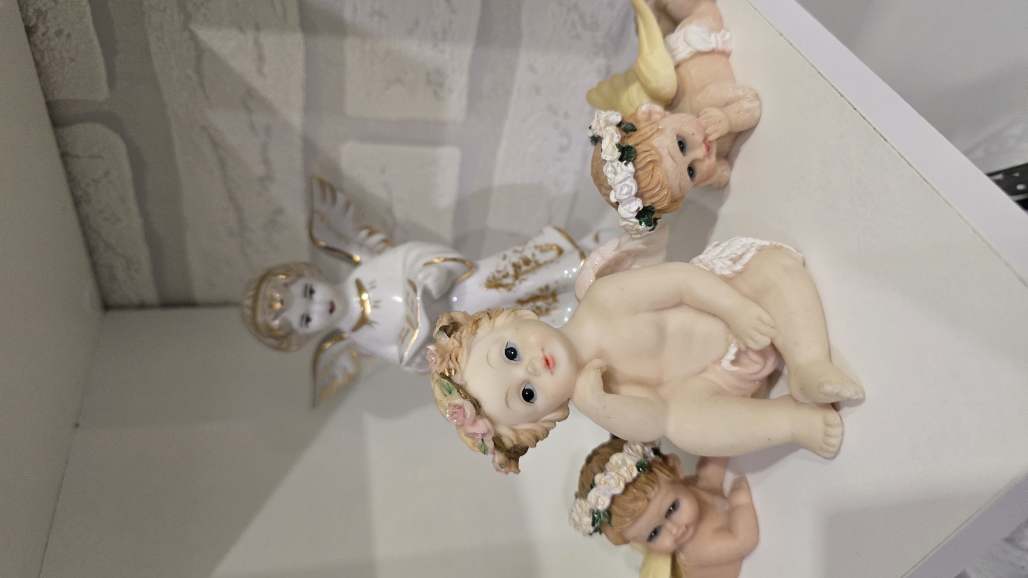 Four porcelain cherub figurines are displayed on a white shelf. The largest figurine is in the center, seated with its left hand resting on its chin and wearing a floral wreath. To the left and right of the central figurine are two smaller cherubs lying down, also wearing floral wreaths. In the background, a taller, standing porcelain angel figure is visible. The background wall appears to be made of white brick. The scene is indoors, likely on a shelf in a home. The location in the city of Верхнеказымский, Russia is not discernible from the image.