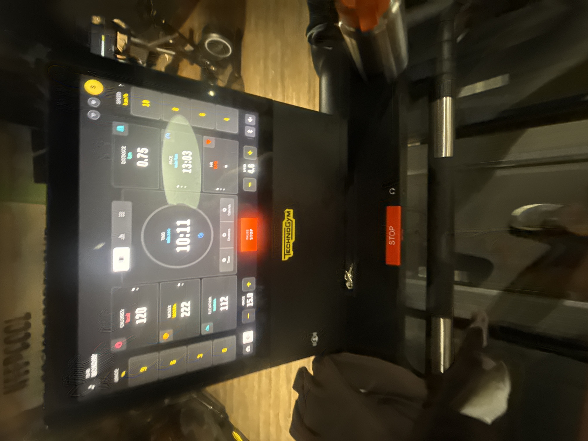 The image provides a close-up, rotated view of a Technogym fitness machine's digital display and controls, likely a treadmill or elliptical, in Paris, France. The screen is actively tracking an exercise session, with several metrics visible against a dark background.
Prominently displayed are:
* "TIME": "10:11" (indicating the duration of the workout)
* "DISTANCE": "0.75 km"
* "PACE": "13:03 min/km"
* "CALORIES": "120"
* "MOVES": "222" (possibly steps or strokes)
* "ELEVATION": "112" (presumably meters or floors climbed)
* "SPEED": "4.6 km/h"
* "GRADE": "15.0" (likely representing an incline percentage)
Several control buttons are visible, including a yellow "S" button in the top left, and two prominent red "STOP" buttons – one labeled "PAUSE STOP" on the screen and another larger physical button on the right side of the machine's console. There are also "+" and "-" buttons for adjusting speed and grade. The "TECHNOGYM" brand logo is visible in yellow text on the console.
The lighting is dim, suggesting an indoor setting, possibly a gym or home workout space. Reflections are noticeable on the screen surface. Part of the machine's handlebars or side rails, made of metal and black material, are visible to the right. A hand, possibly wearing a gray glove, is visible in the lower right foreground, resting on the machine's surface. Another object, possibly a water bottle or cup, is partially visible in the upper right. The text "NEEPCOOL" is seen upside down on a surface in the bottom left, suggesting a brand name on a nearby item.
